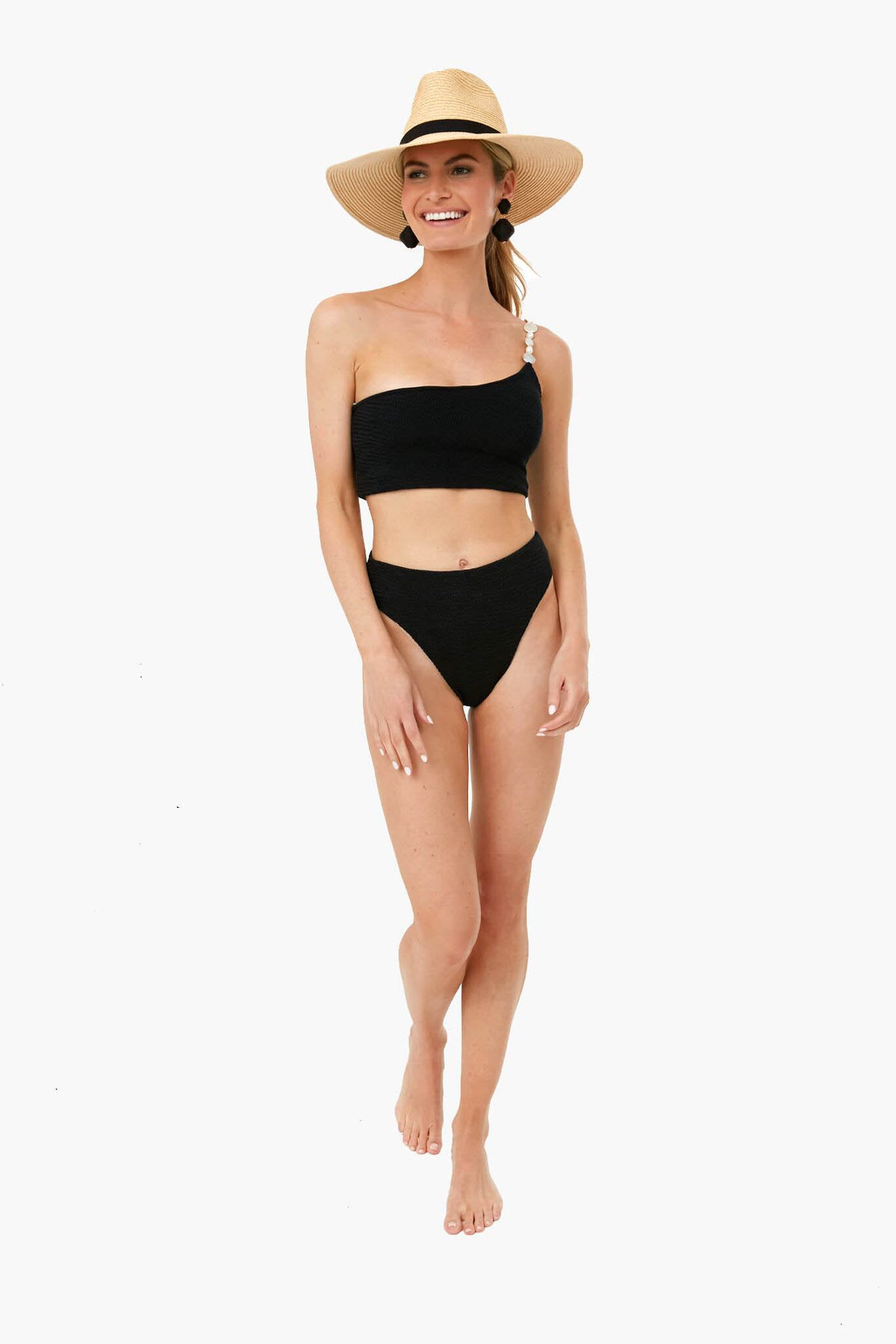 A woman stands barefoot, smiling, in a Maygel Coronel black one-shoulder bikini with a pearl strap and a wide-brimmed straw hat.
