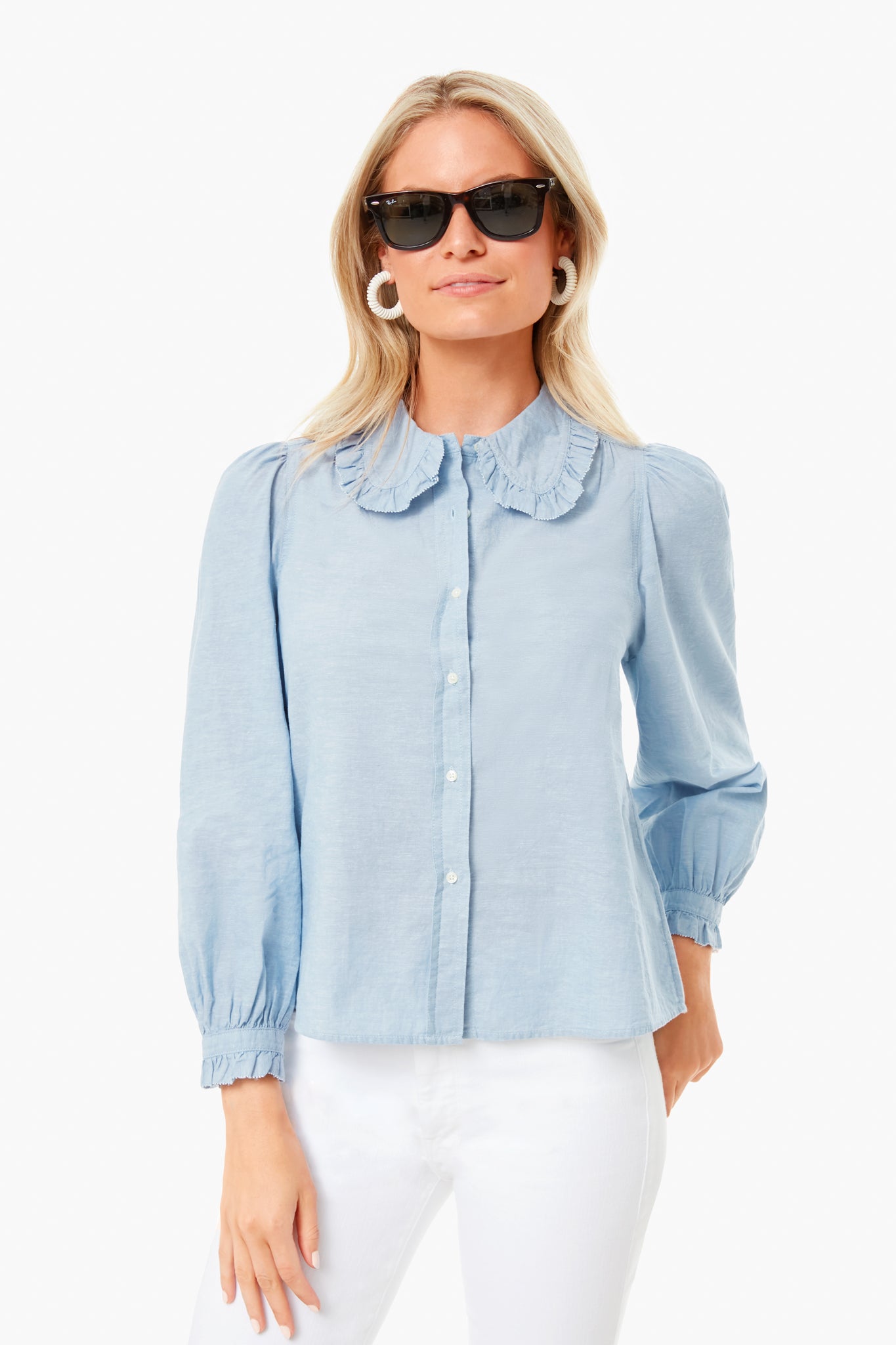 A woman stands confidently in black sunglasses, white pants, and THE GREAT.'s light blue Hemingway Top featuring a ruffled collar.
