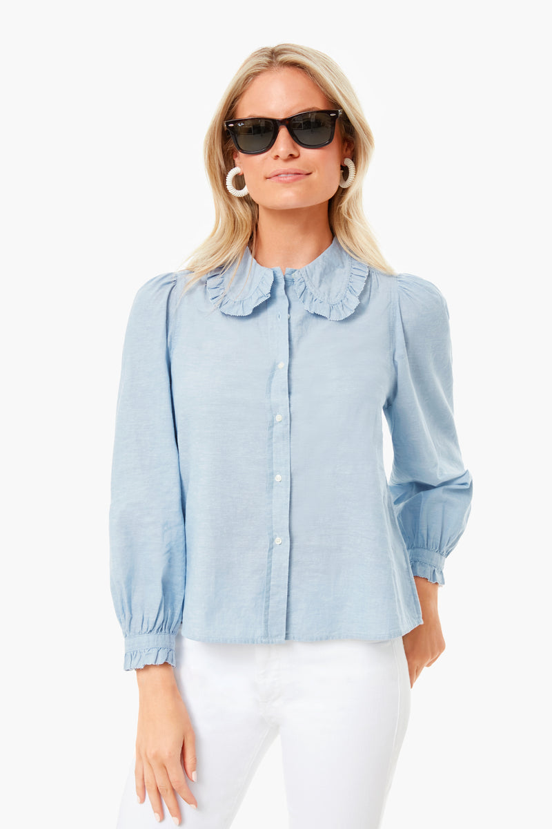 A woman stands confidently in black sunglasses, white pants, and THE GREAT.'s light blue Hemingway Top featuring a ruffled collar.