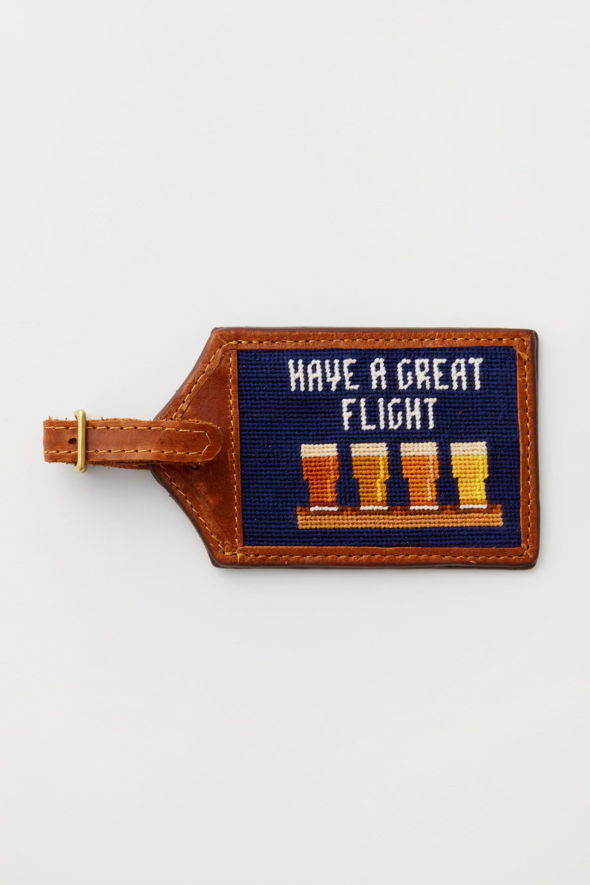 The Smathers and Branson brown leather luggage tag features hand-stitched needlepoint with "Have a Great Flight" above four beer glasses.