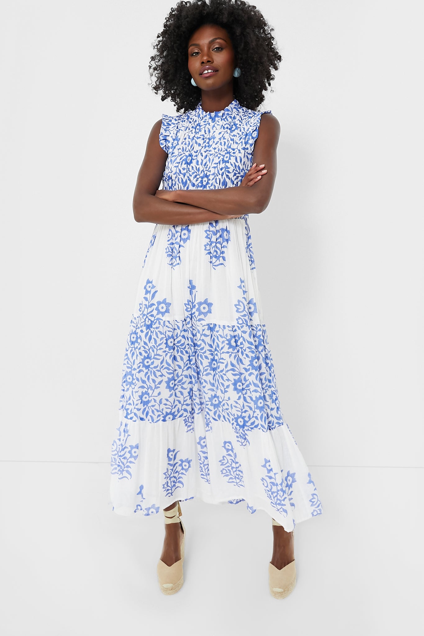 A woman in an Oliphant sleeveless smocked maxi cotton voile dress with blue floral patterns, arms crossed, exudes effortless, vacation-ready style.