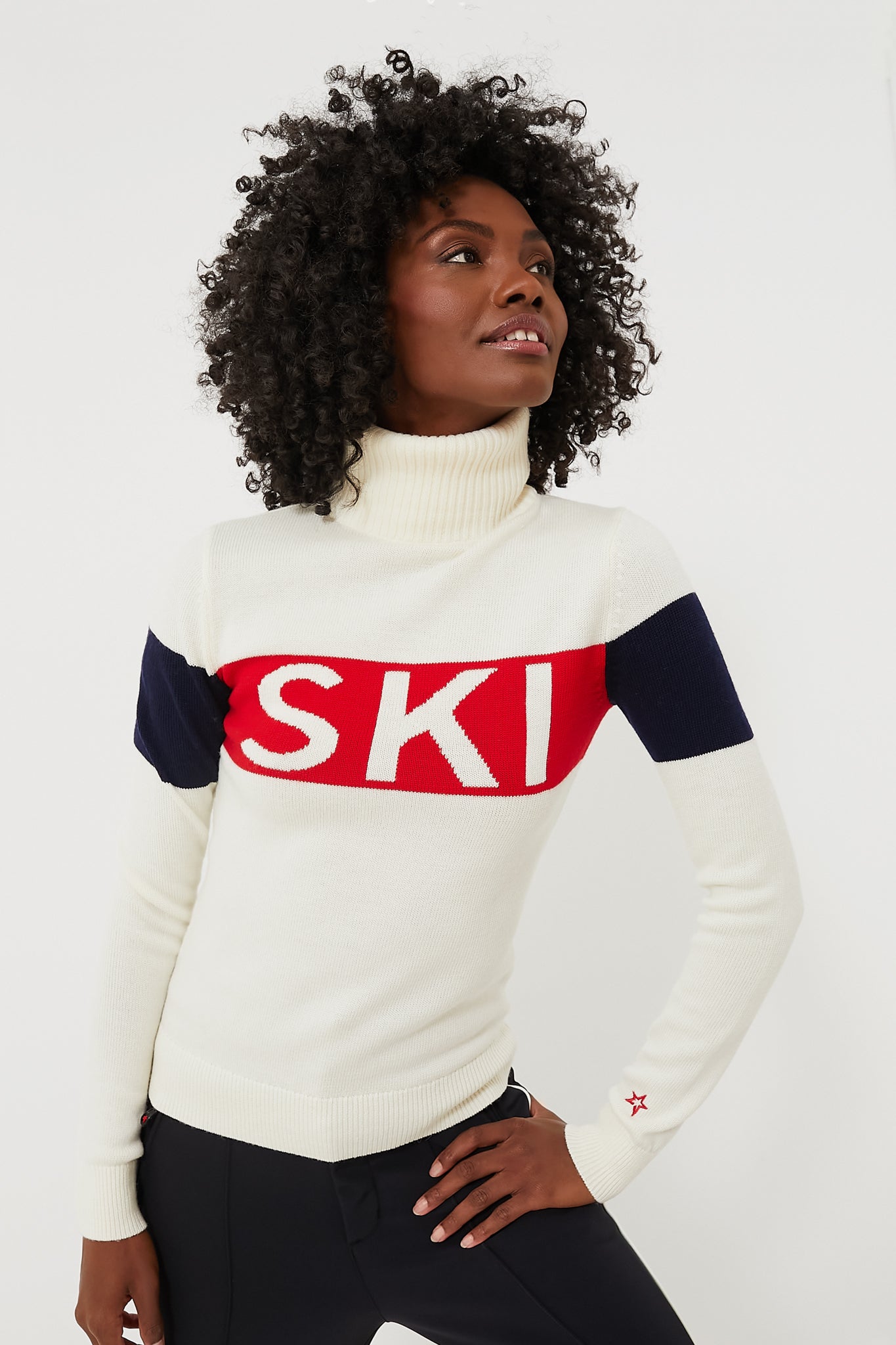 A woman wears a Perfect Moment ski sweater with a white turtleneck, dark sleeves, and a bold red and white SKI logo across the chest.