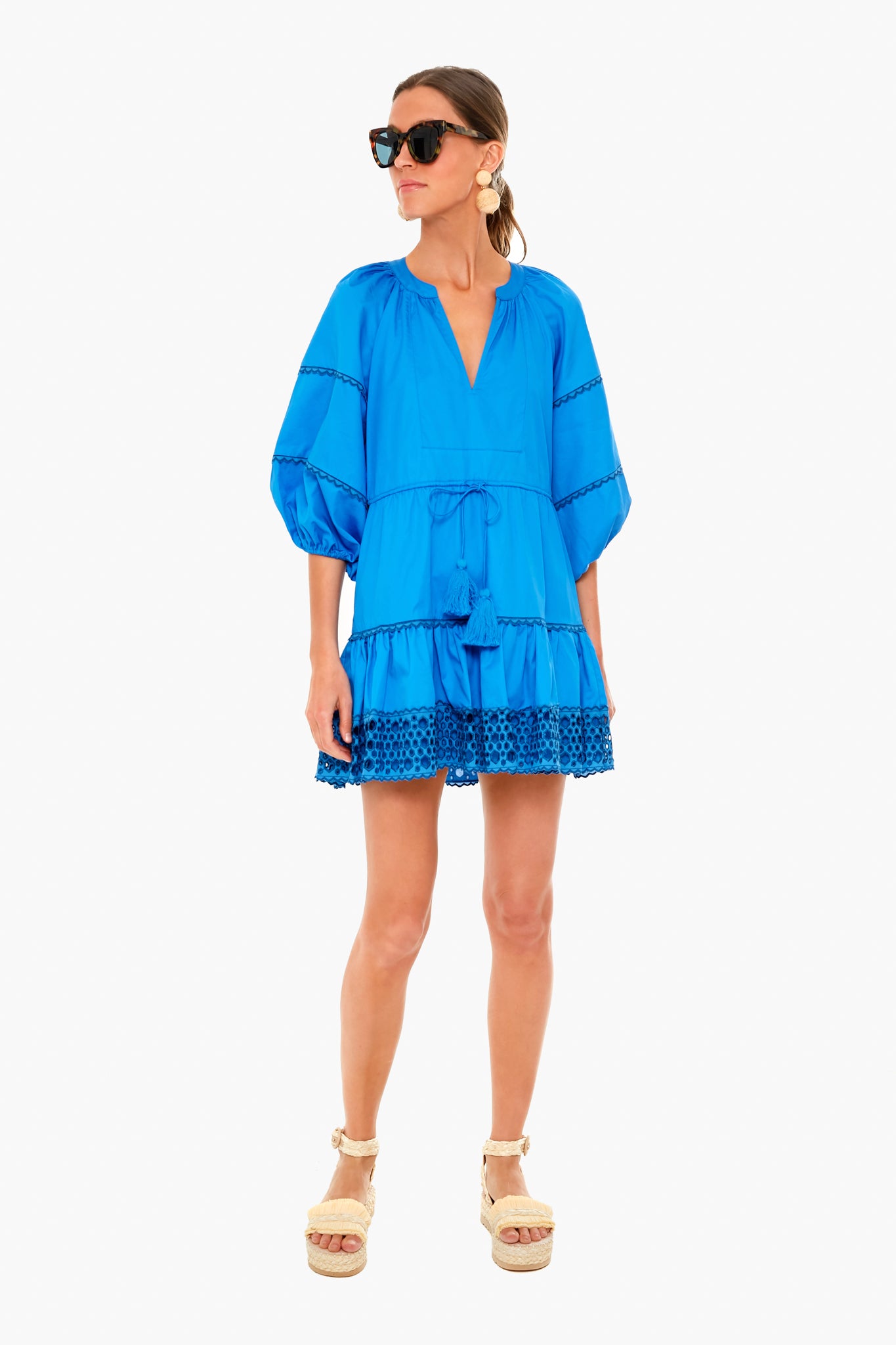 A woman wears a vibrant blue ALEXIS dress with puffed sleeves, lace trim, waist tie, styled with platform sandals and sunglasses.