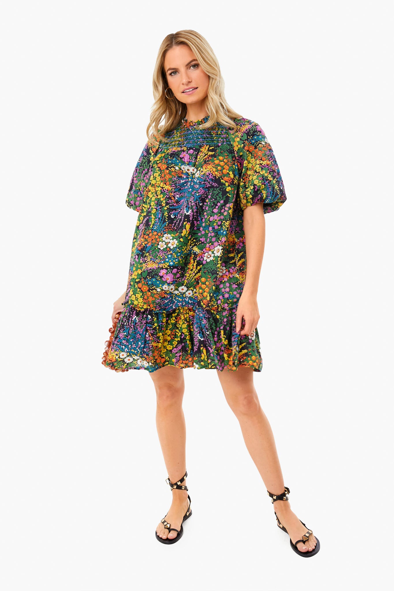 A woman wears a Banjanan organic cotton puff sleeve mini dress with a colorful floral print and ruffled hem, paired with black sandals.