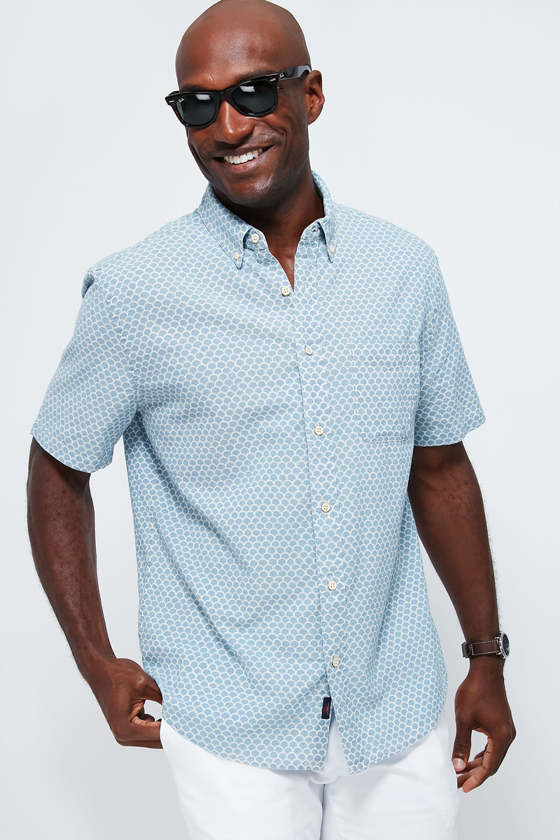 A man smiles confidently wearing Faherty sunglasses, a light blue patterned short-sleeve stretch Playa shirt, and white pants.