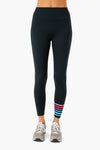 A person wears Addison Bay black high-rise activewear leggings with colorful stripes near the left ankle and gray sneakers, shown from waist to feet.