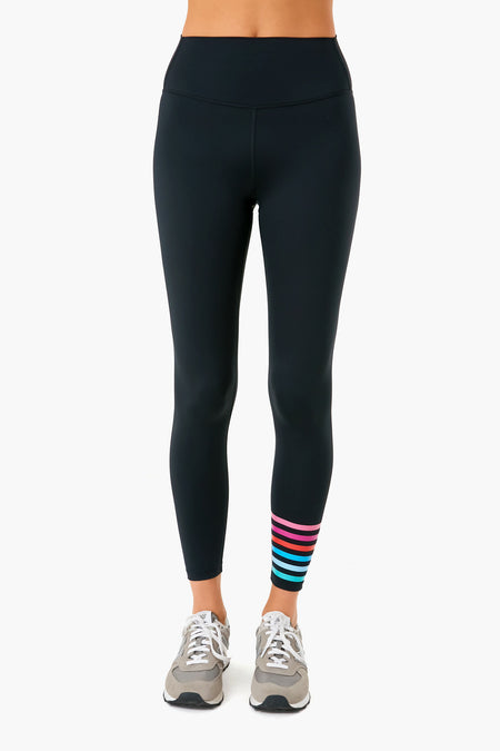 A person wears Addison Bay black high-rise activewear leggings with colorful stripes near the left ankle and gray sneakers, shown from waist to feet.
