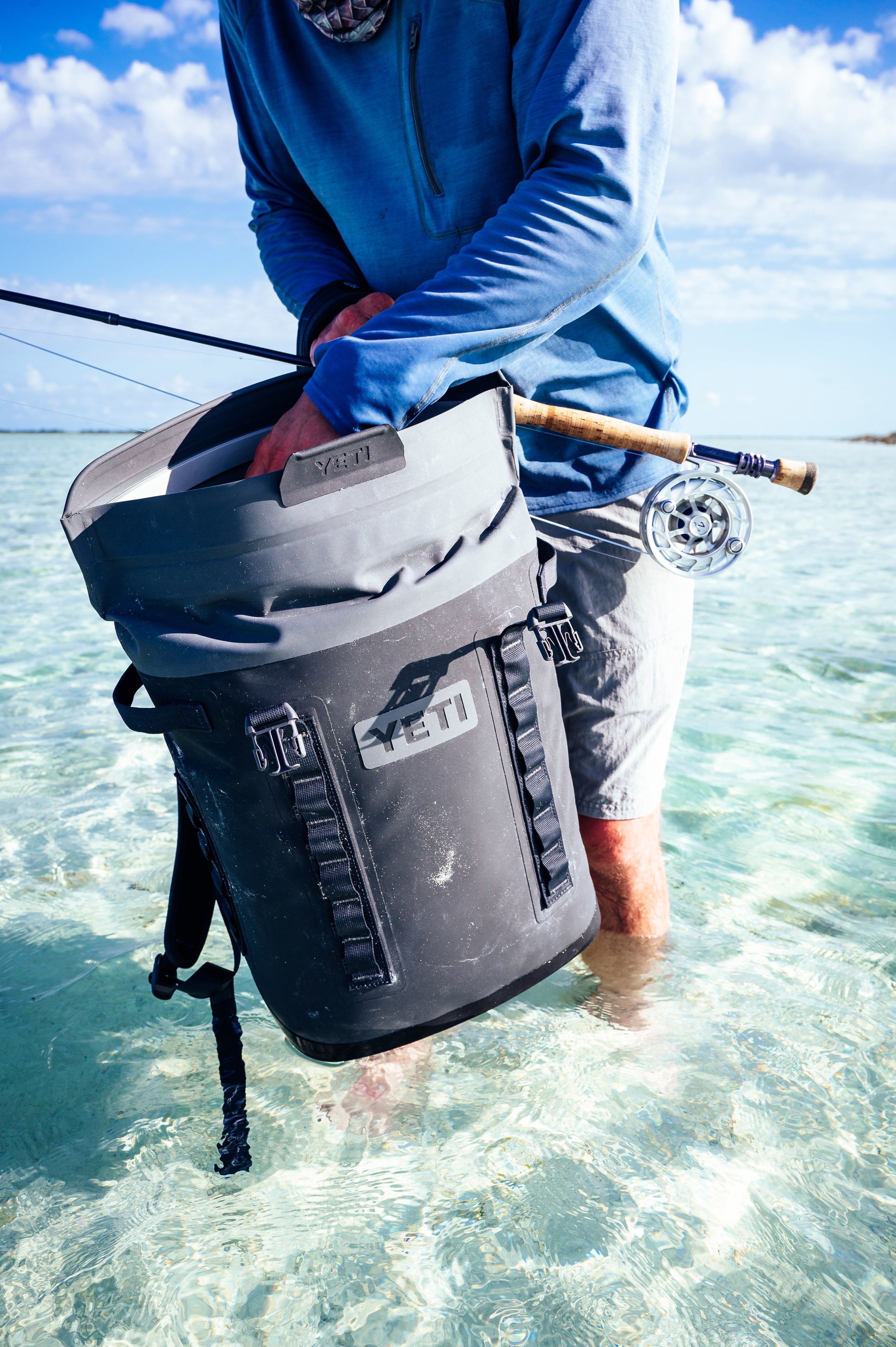Person standing in water holding a fly-fishing rod and a classic Navy Hopper M20 Backpack, embodying effortless adventure and fun with high-end YETI quality.