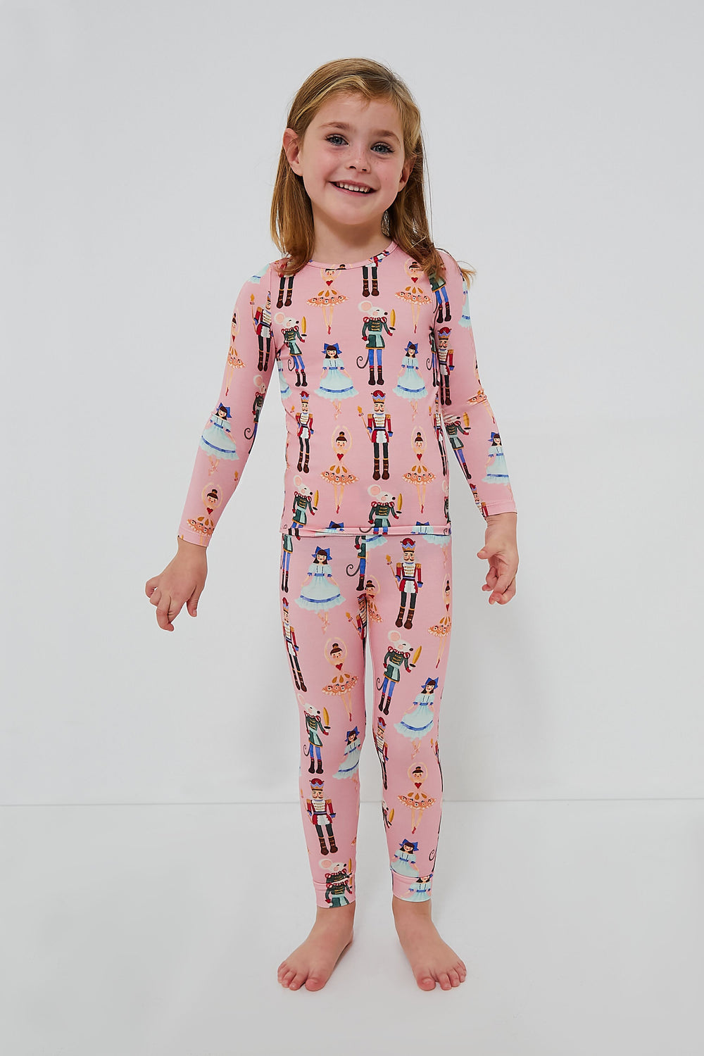 A young child stands barefoot in a Pink Chicken Kids Bamboo PJ Set featuring a playful Toy Nutcracker print.