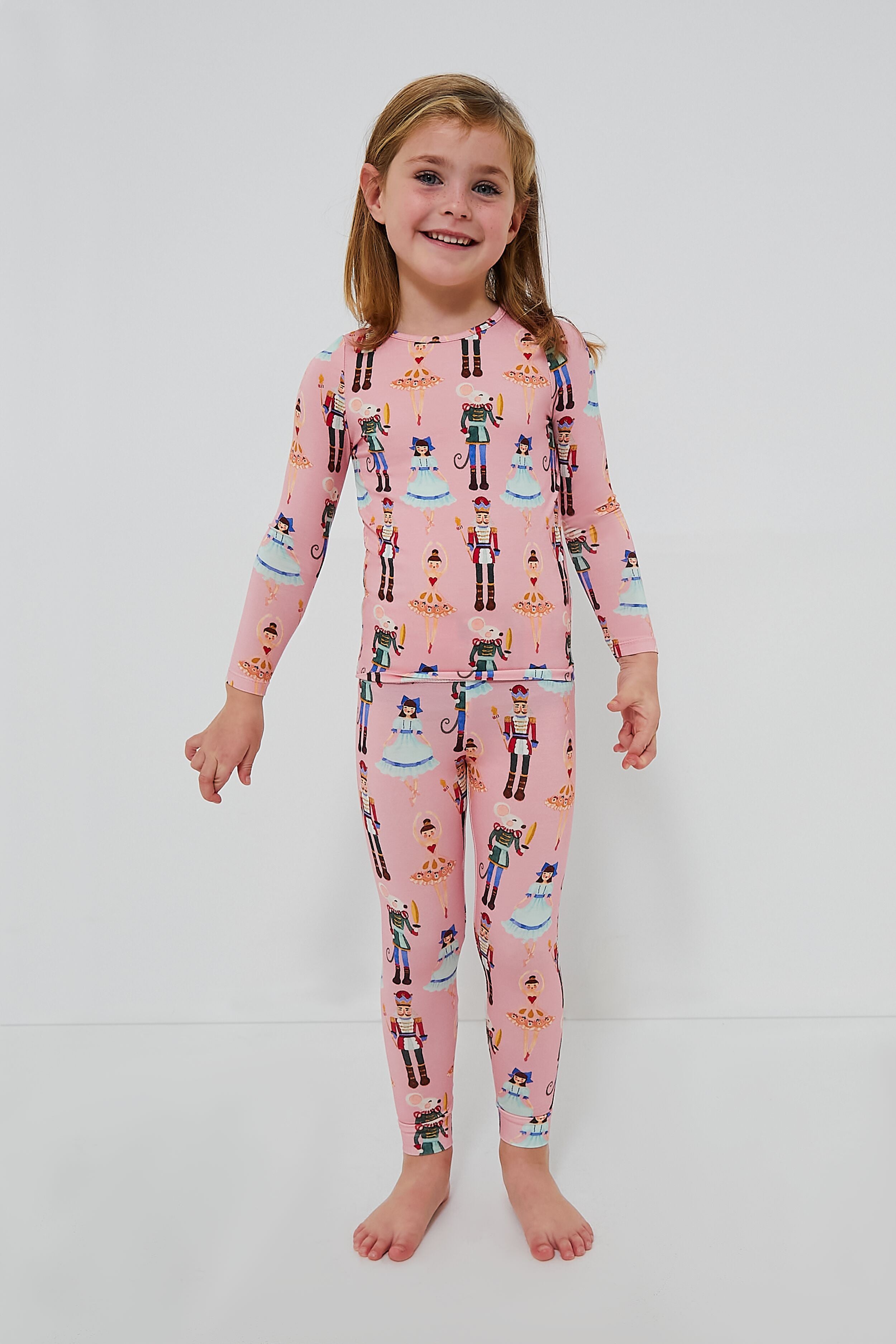 A young child stands barefoot in a Pink Chicken Kids Bamboo PJ Set featuring a playful Toy Nutcracker print.