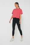 A woman wears a Tnuck Sport pink cropped boyfriend tee with moisture-wicking fabric, black leggings, and white sneakers.