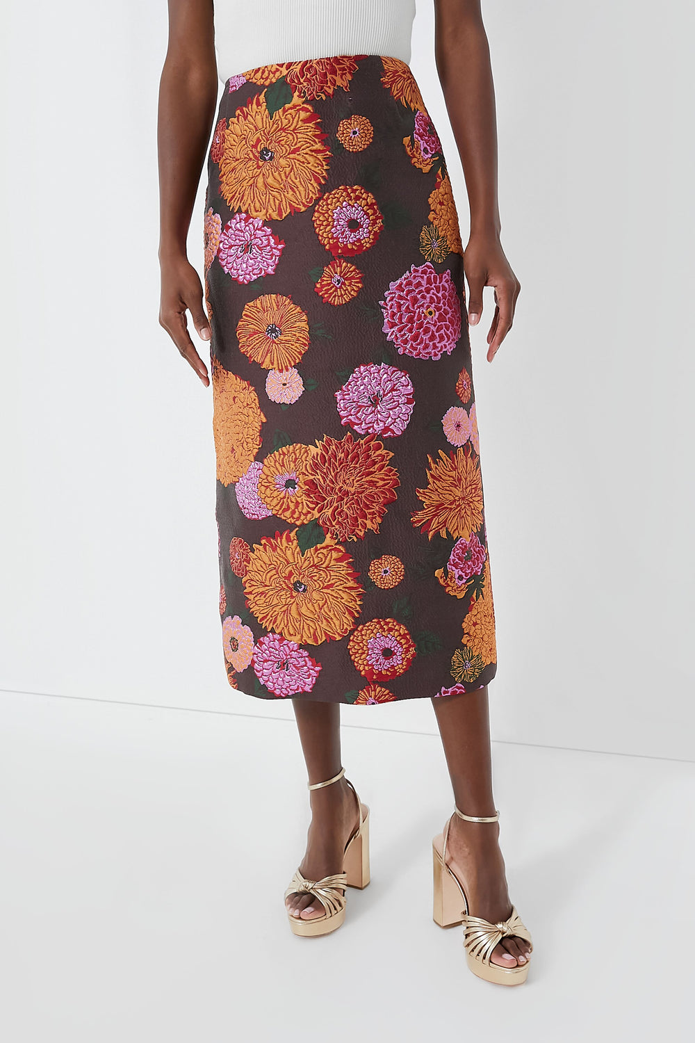 A person wears a white top, a long dark floral La DoubleJ skirt with orange and pink flowers—an elegant Milanese-inspired look—and gold high-heeled sandals.