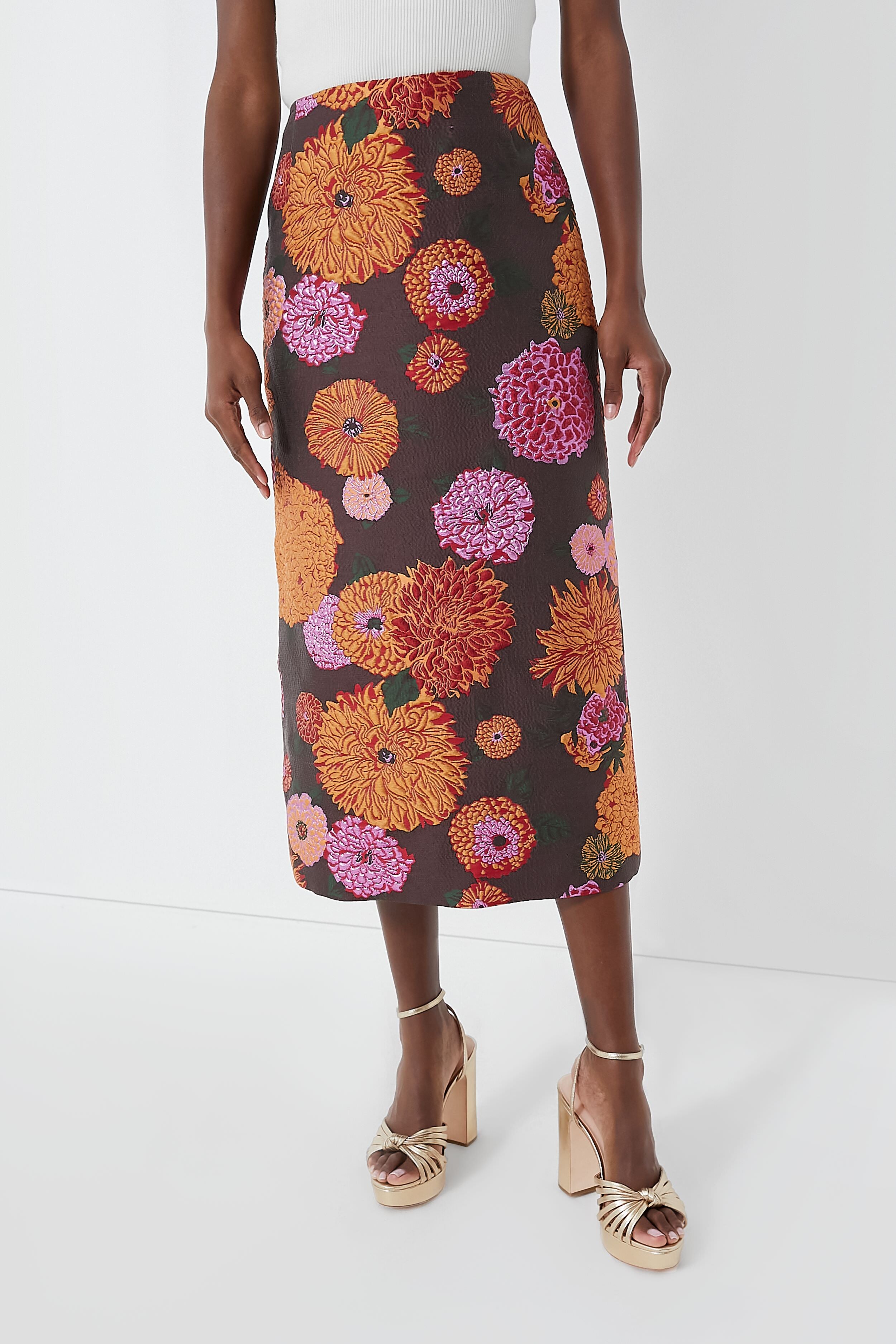 A person wears a white top, a long dark floral La DoubleJ skirt with orange and pink flowers—an elegant Milanese-inspired look—and gold high-heeled sandals.