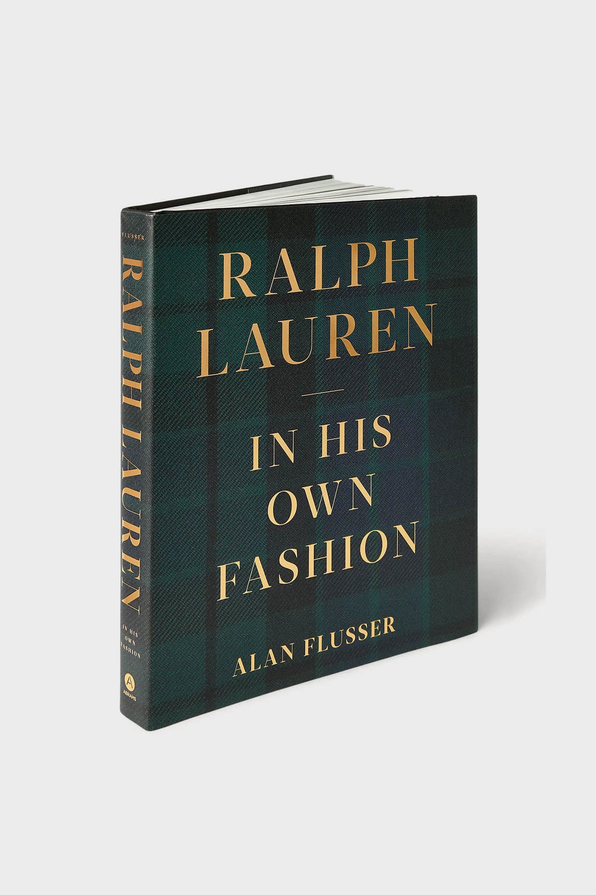 Hardcover book titled "Ralph Lauren: In His Own Fashion" by Alan Flusser, published by Abrams Books with gold text on a dark plaid cover—an essential American fashion biography.