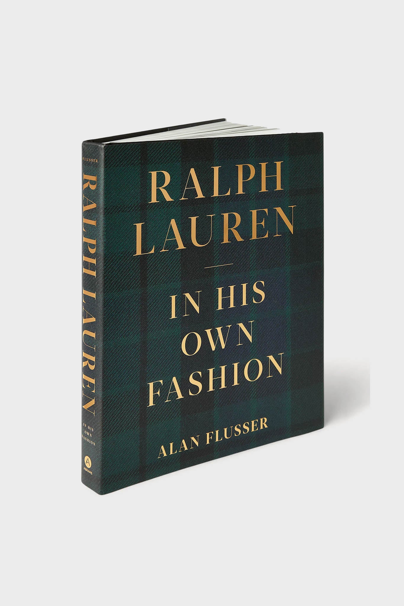 Hardcover book titled "Ralph Lauren: In His Own Fashion" by Alan Flusser, published by Abrams Books with gold text on a dark plaid cover—an essential American fashion biography.