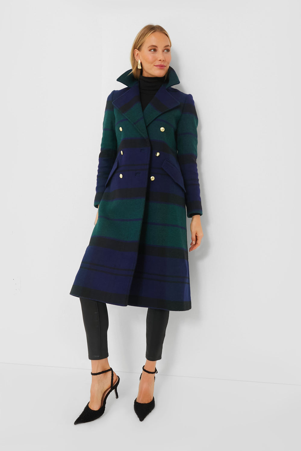A woman wears a long, double-breasted Barbour blue and green tartan plaid wool blend coat over black pants and a black turtleneck, paired with black heels.
