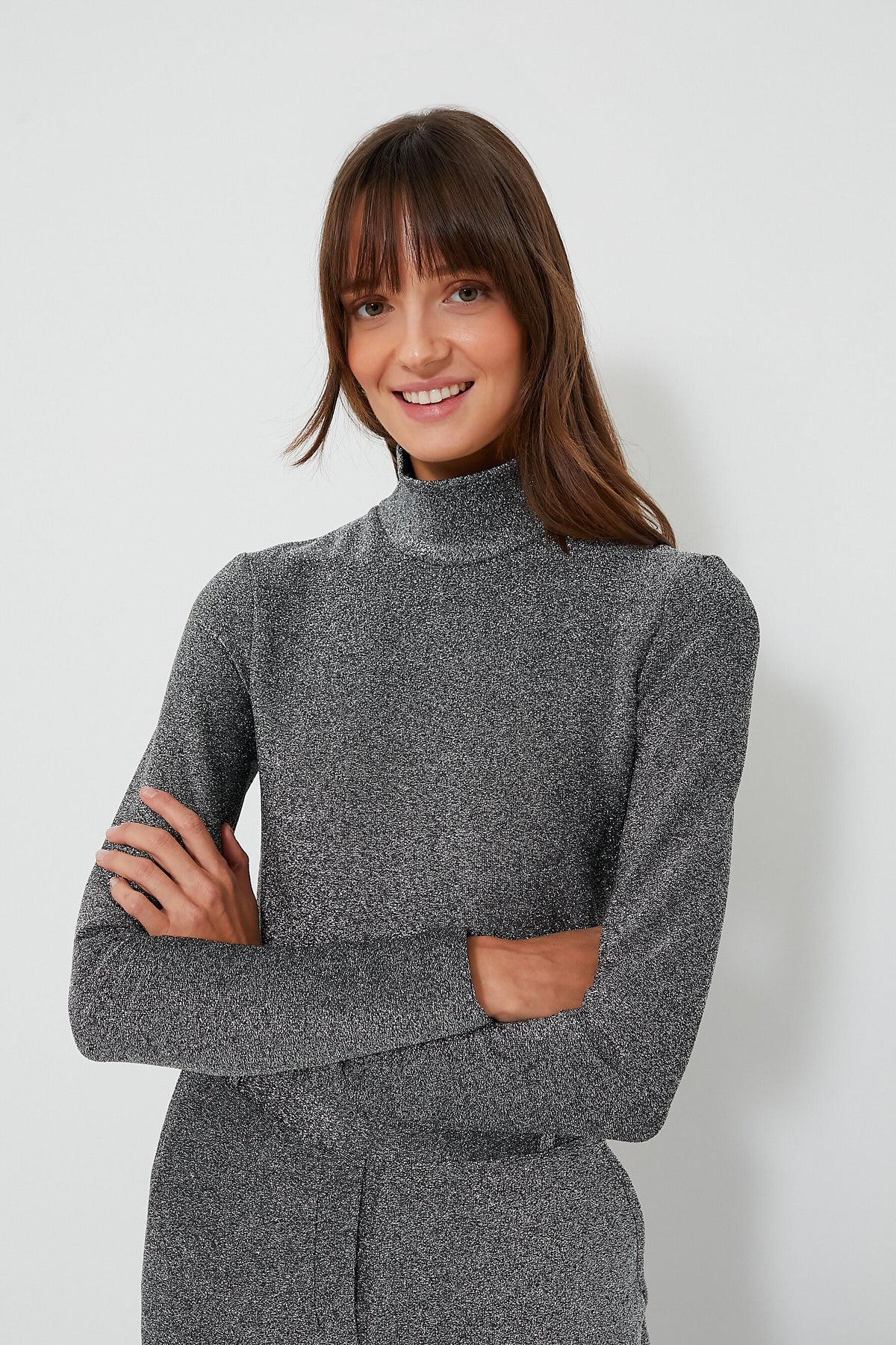 A woman with long brown hair and bangs, smiling with arms crossed, wears a gray fitted Baum Und Pferdgarten women’s designer turtleneck top.