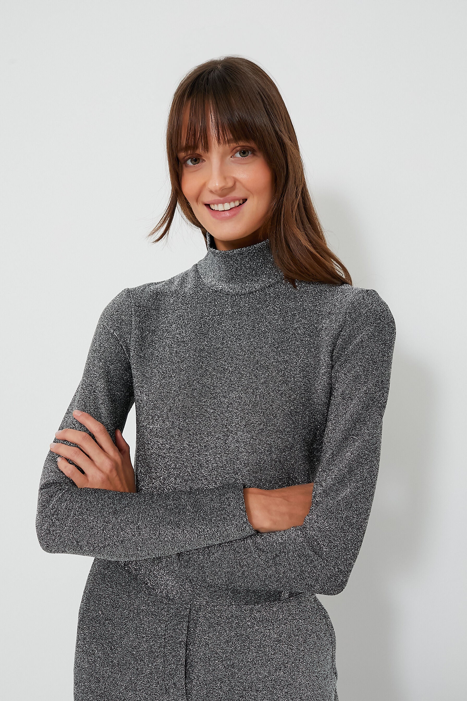 A woman with long brown hair and bangs, smiling with arms crossed, wears a gray fitted Baum Und Pferdgarten women’s designer turtleneck top.