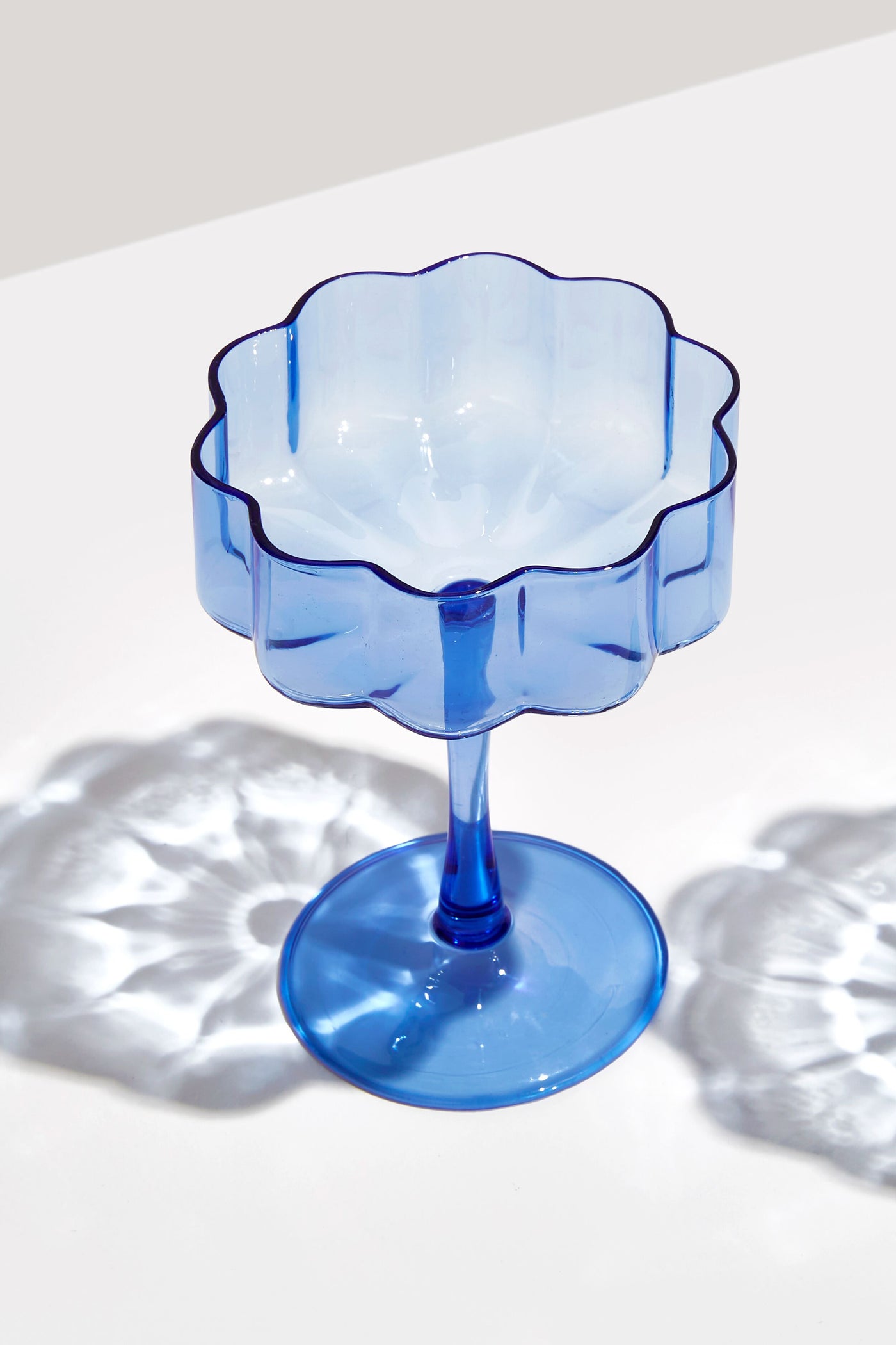 FAZEEK Blue Wave coupe dessert cup with a scalloped edge, made from hand-blown borosilicate glass, rests on a white surface casting delicate light and shadow patterns.