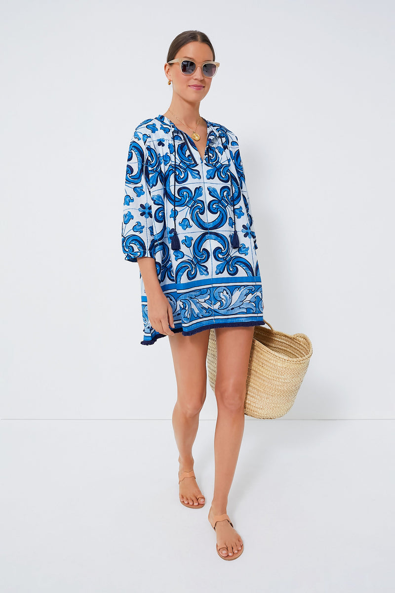 A woman in sunglasses and sandals wears the Tnuck Beach Capri Blue Majolica Tile Bondi Coverup and holds a woven tote bag while standing.