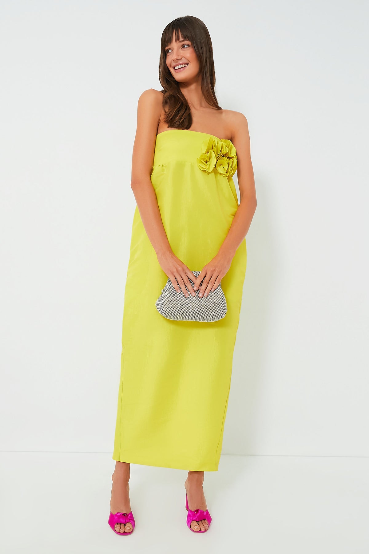 A woman wears a strapless yellow KIKA VARGAS Rossetta Dress with floral detail, holds a silver clutch, and pairs it with pink high heels.