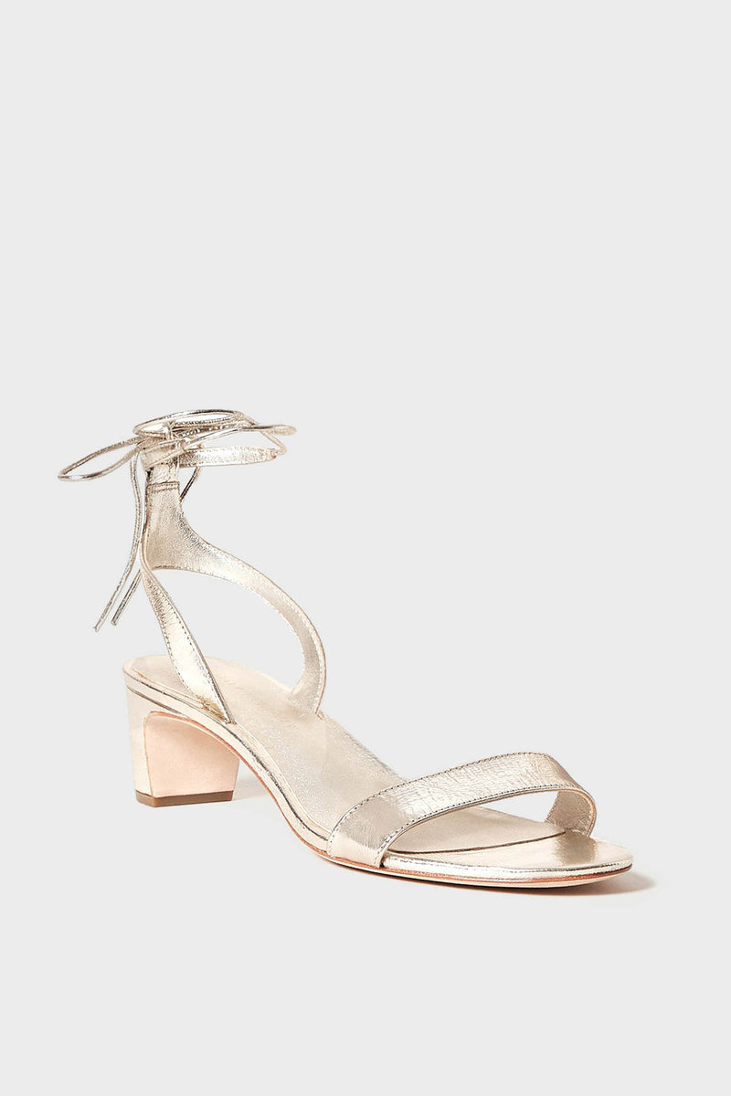 Loeffler Randall's metallic leather ankle wrap sandal features a low block heel and open toe design, showcased in a minimalist setting.
