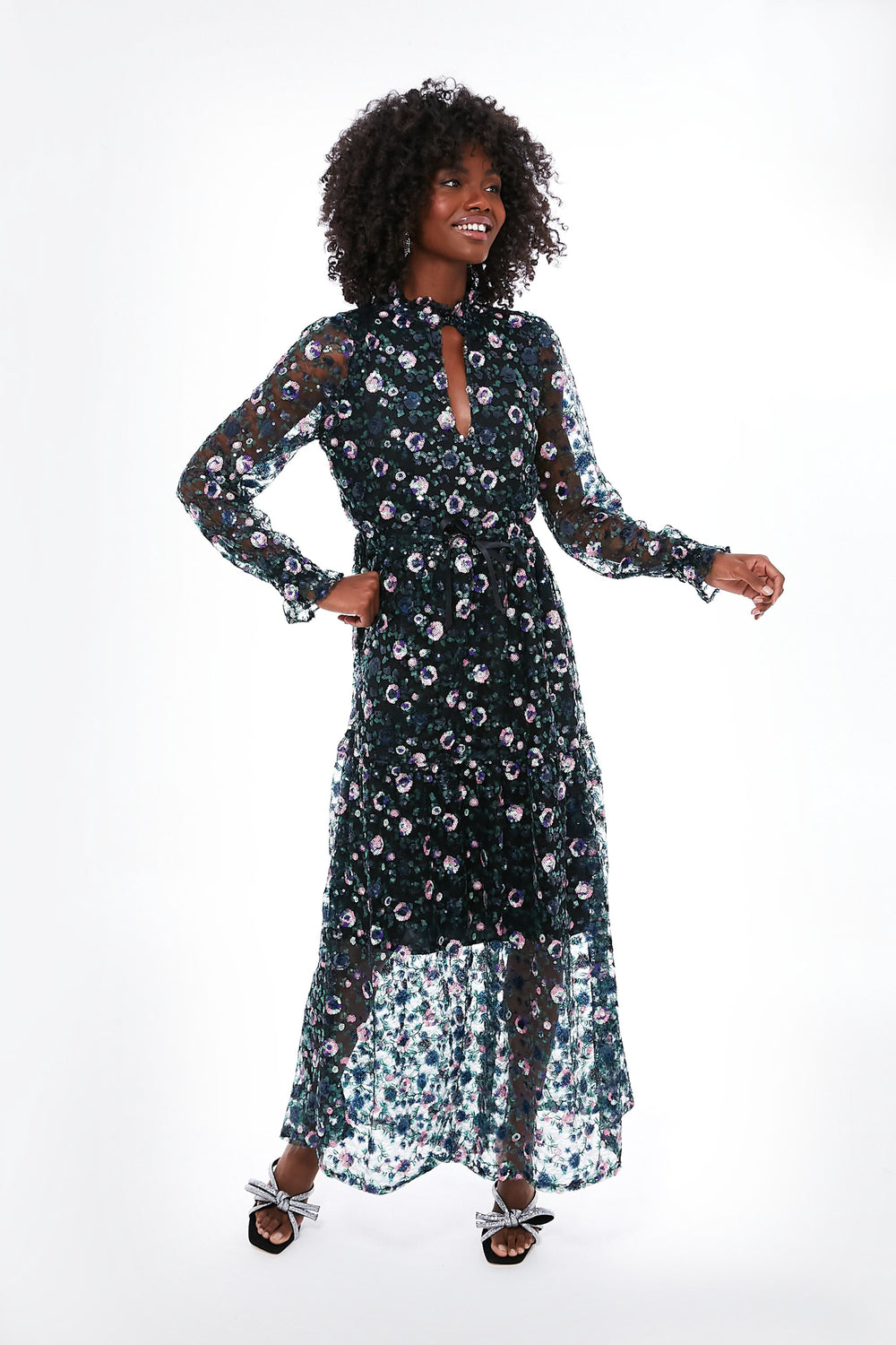 A woman models the elegant Emerson Fry Floral Lucybella Dress—a long-sleeved, sheer maxi with floral pattern—styled with a black belt and silver heeled sandals.