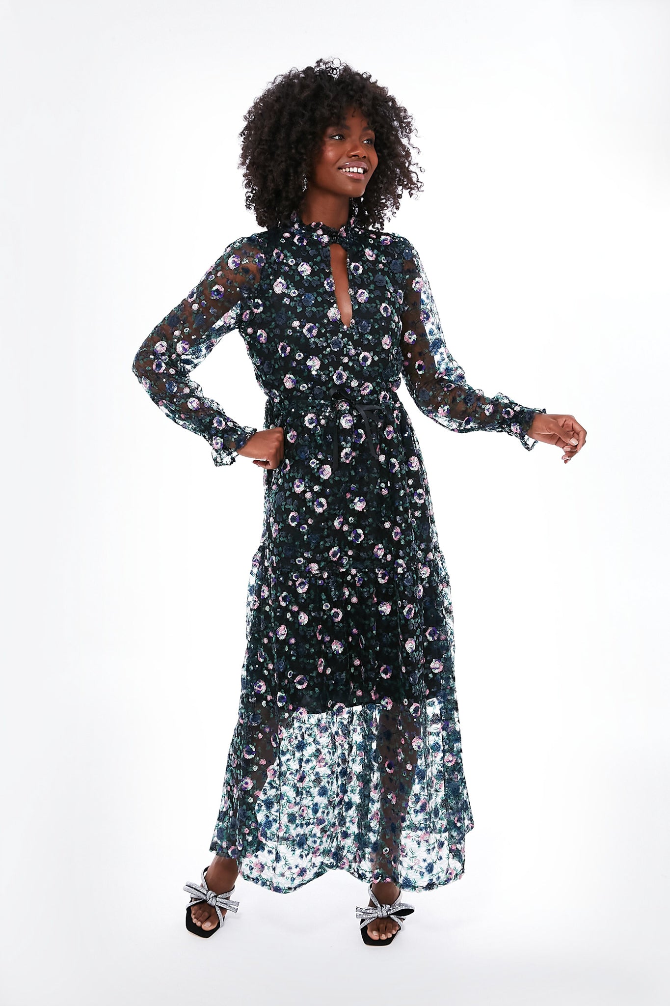 A woman models the elegant Emerson Fry Floral Lucybella Dress—a long-sleeved, sheer maxi with floral pattern—styled with a black belt and silver heeled sandals.
