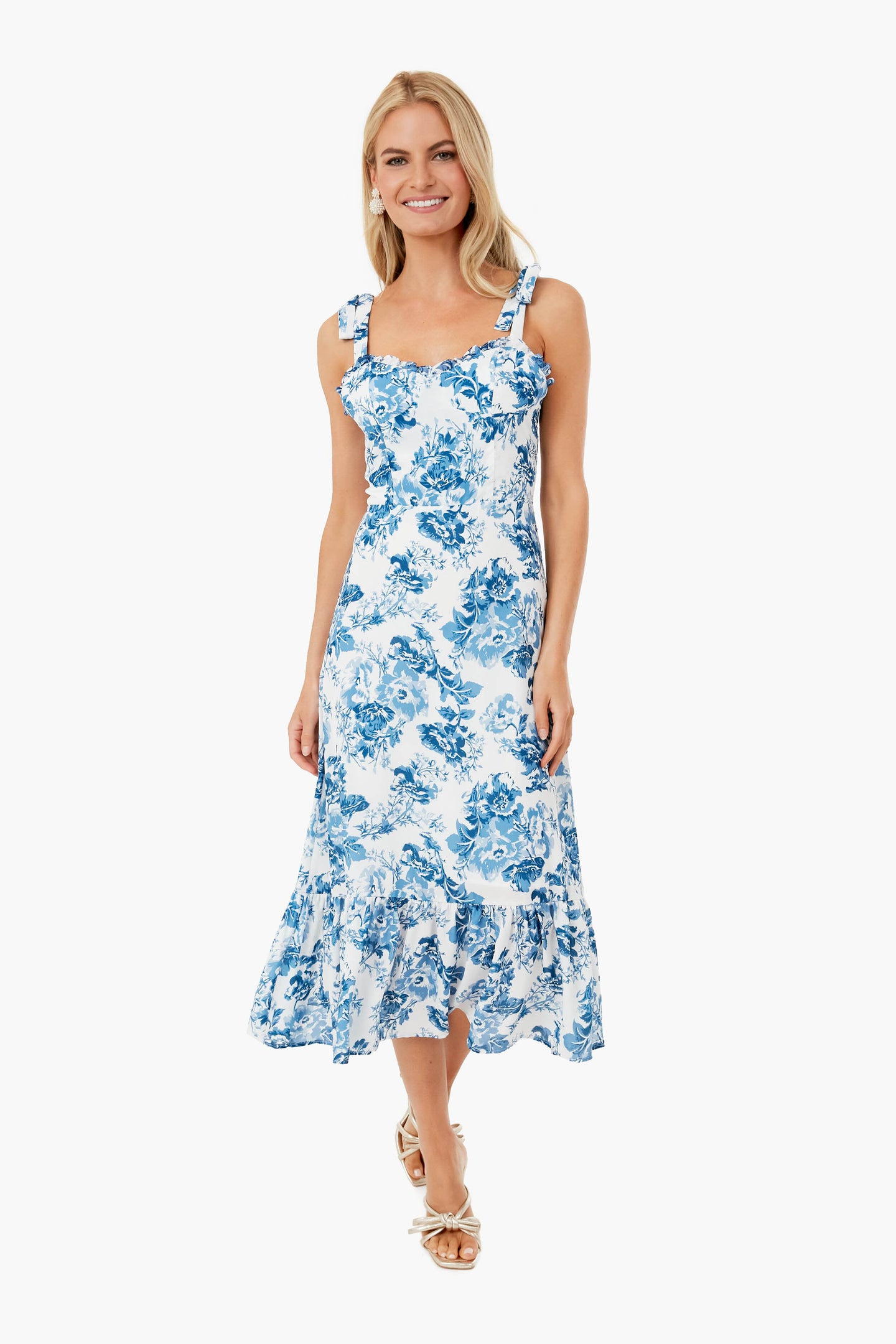 A woman stands and smiles in a white setting, wearing an O.P.T blue floral midi dress with shoulder straps.