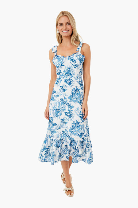 A woman stands and smiles in a white setting, wearing an O.P.T blue floral midi dress with shoulder straps.