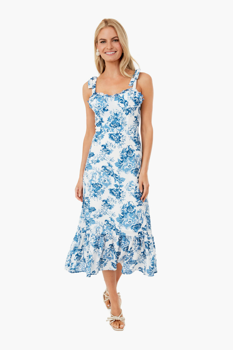 A woman stands and smiles in a white setting, wearing an O.P.T blue floral midi dress with shoulder straps.