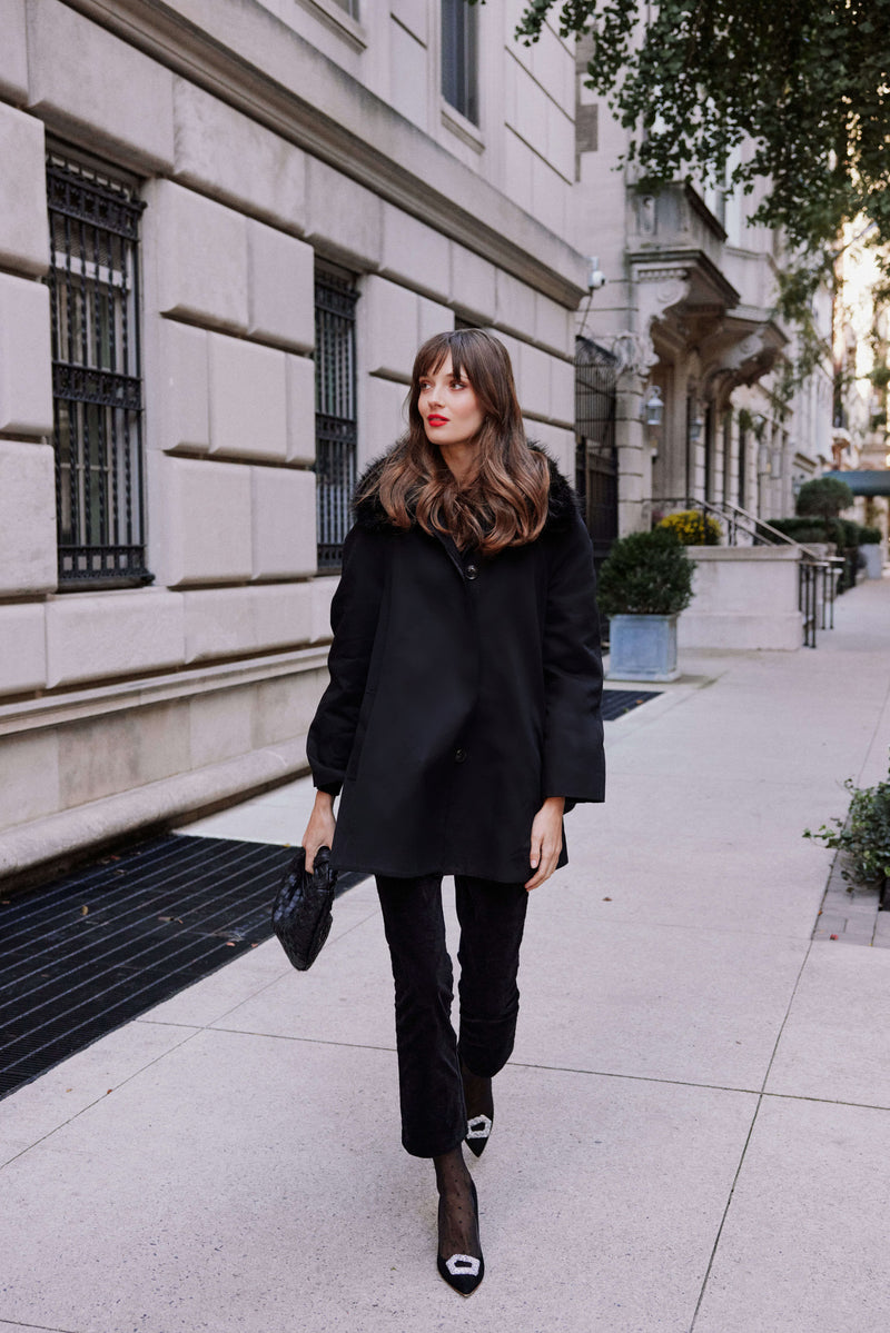 A woman in a Tuckernuck black coat, high-waisted velvet pants, and black heels walks along a city sidewalk by a beige stone building with iron window bars.