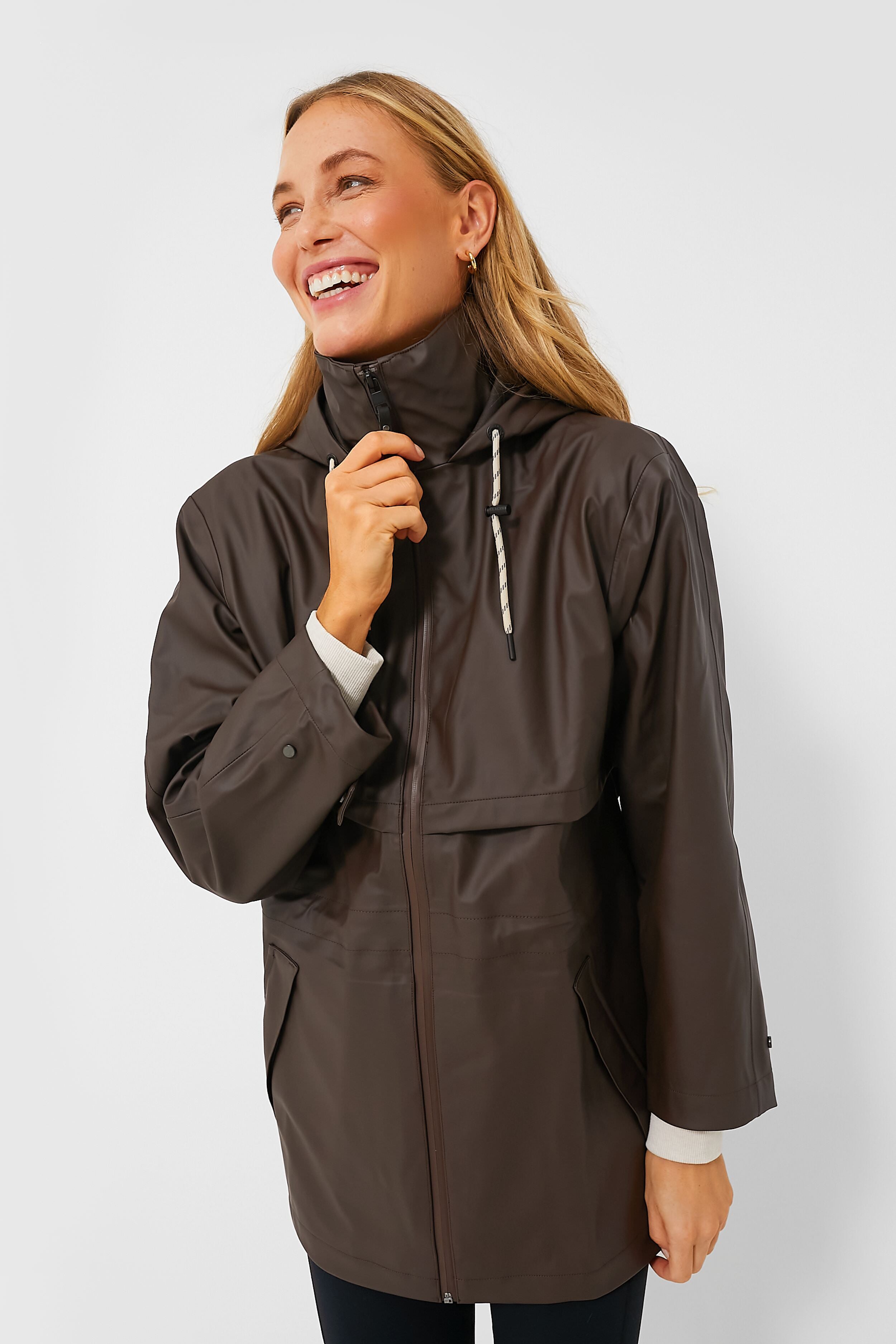 A woman wearing a Varley Alyssa Rain Jacket, an oversized women's raincoat in dark brown, smiles and holds the collar.