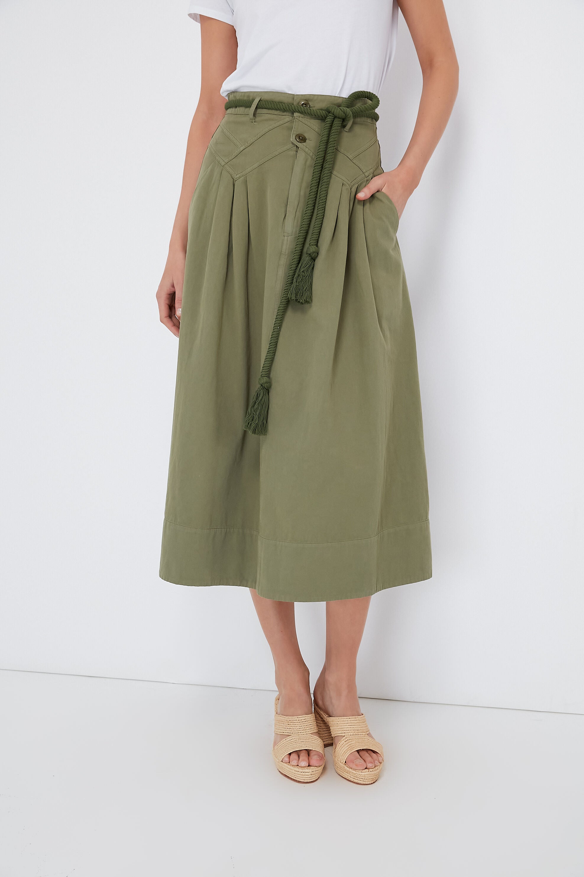 A person wears a white t-shirt, THE GREAT. olive green Army Field Skirt with a rope belt in soft garment-dyed twill, and beige sandals.