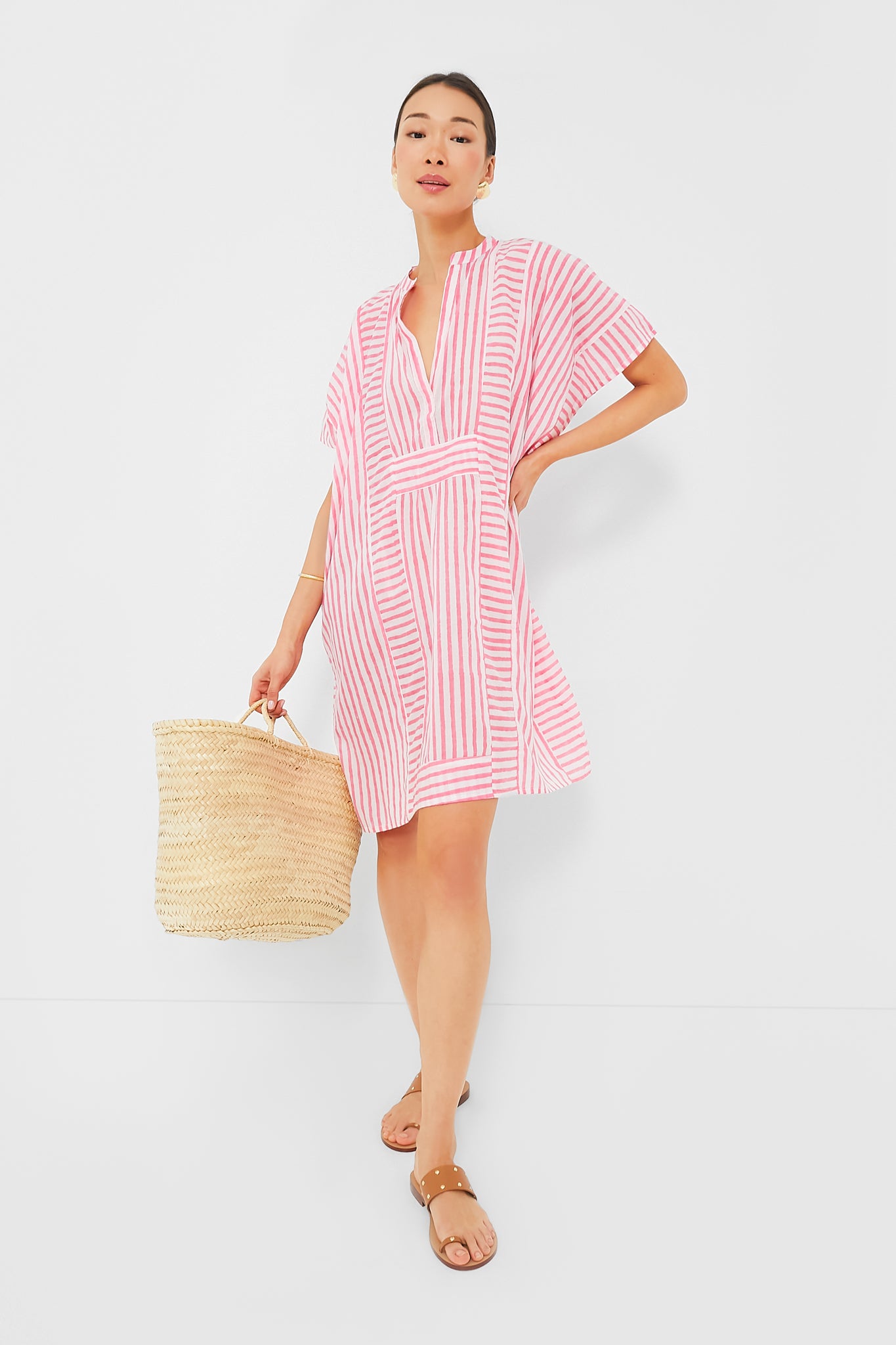 A woman wears a pink and white striped short dress from Love Tomas by Emerson Fry, tan sandals, and carries a woven straw bag.