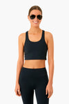 A woman in black sunglasses, an Addison Bay Black Arch Sports Bra, and matching black leggings stands smiling with her hands by her sides.