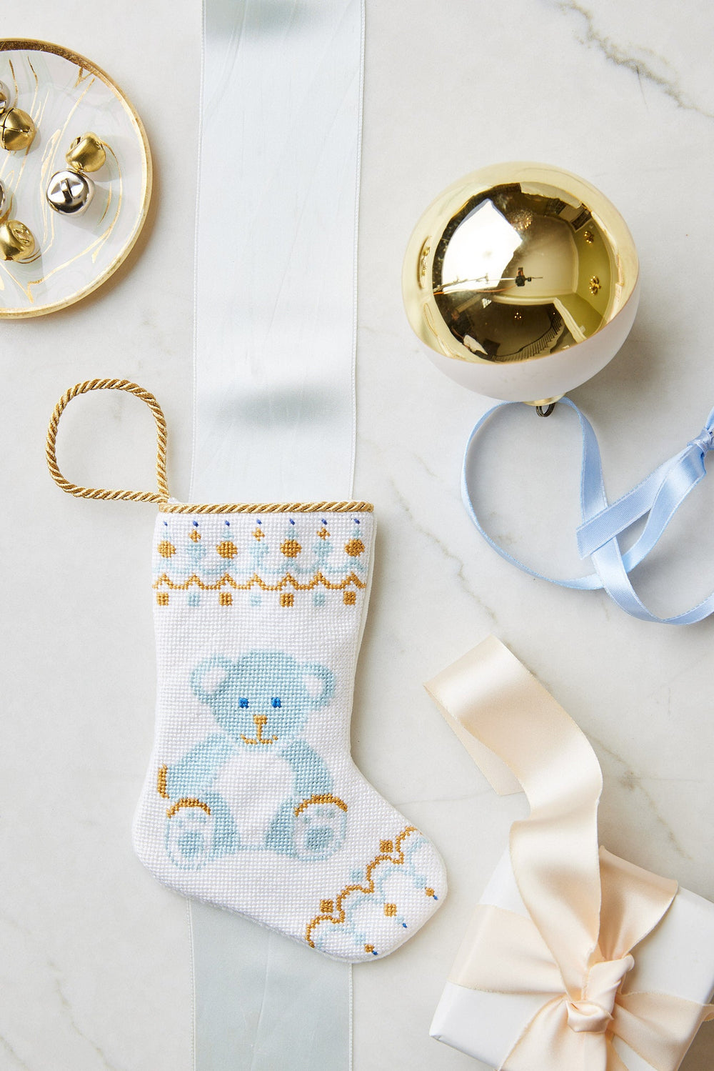 A small hand-stitched Bauble Stockings Christmas stocking with a blue teddy bear design is displayed on marble with a gold ornament, branded ribbon, bells, and a gift box.