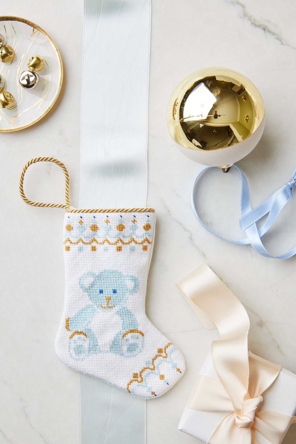 A small hand-stitched Bauble Stockings Christmas stocking with a blue teddy bear design is displayed on marble with a gold ornament, branded ribbon, bells, and a gift box.