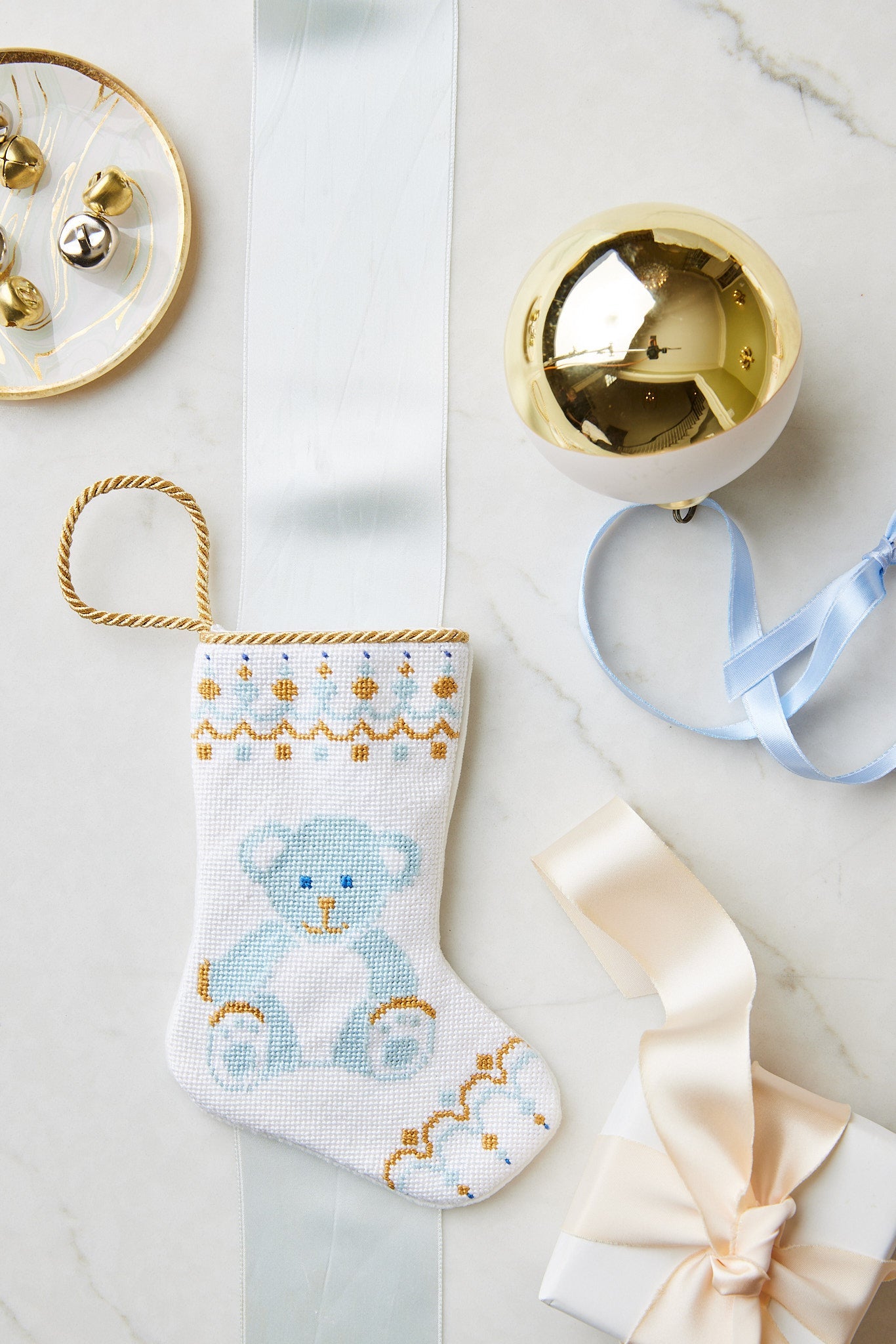 A small hand-stitched Bauble Stockings Christmas stocking with a blue teddy bear design is displayed on marble with a gold ornament, branded ribbon, bells, and a gift box.