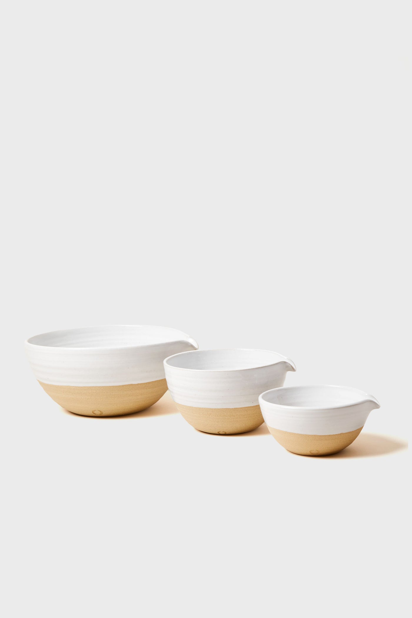 Three ceramic Pantry Bowls from Farmhouse Pottery, Inc. feature white tops and natural clay bottoms in varying sizes, arranged in a row—an ideal accent for any farmhouse kitchen.