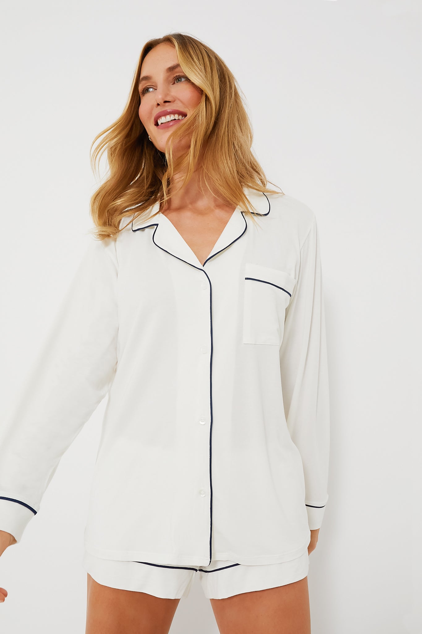 Wearing the Eberjey Ivory Gisele PJ Set—white long-sleeve top and matching shorts with black piping—a person smiles and looks to the side, highlighting cozy TENCEL Modal pajamas.