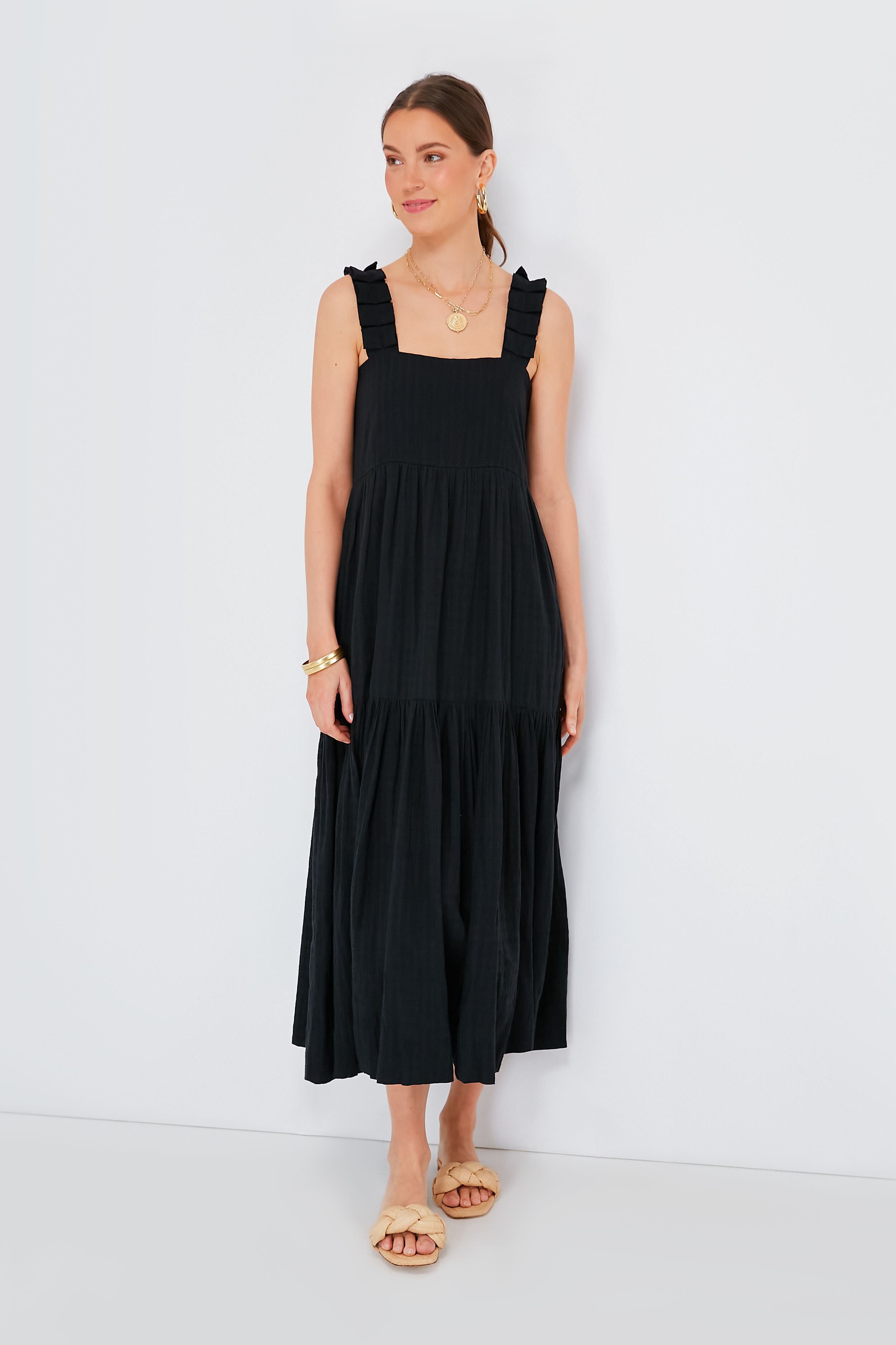 A woman wears MIRTH’s sleeveless, black ankle-length artisan dress with ruffled straps, paired with gold jewelry and beige sandals—an elegant example of ethical resortwear.
