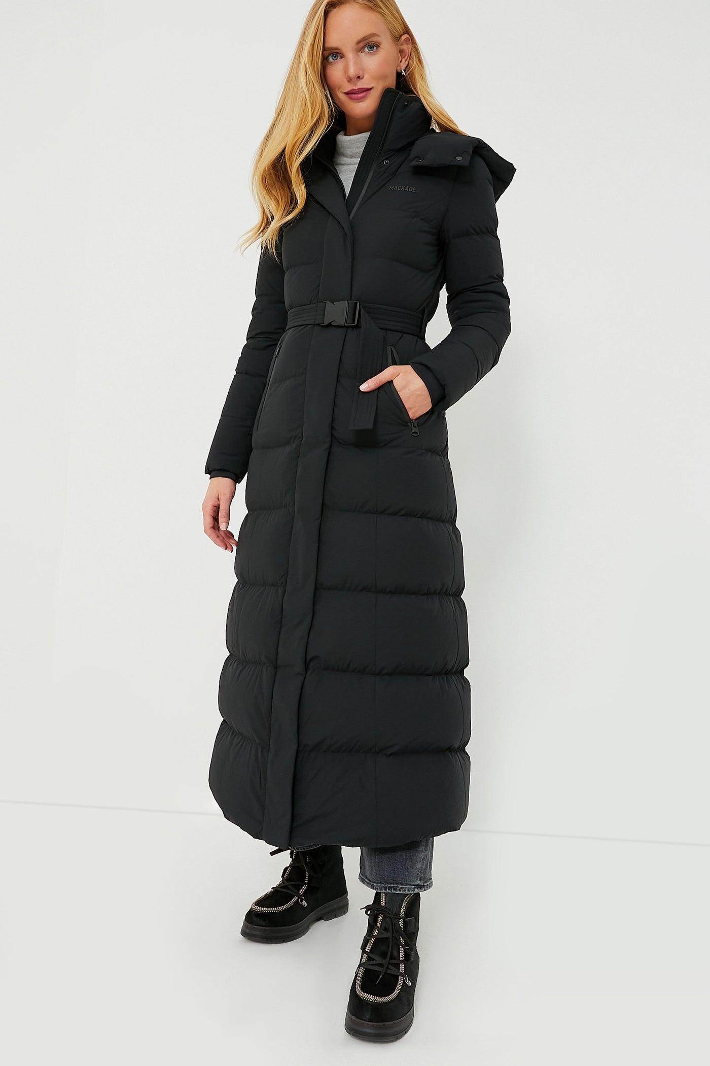A person wears a MACKAGE women's winter parka—a long black puffer coat with a belt—styled with black boots and gray jeans.