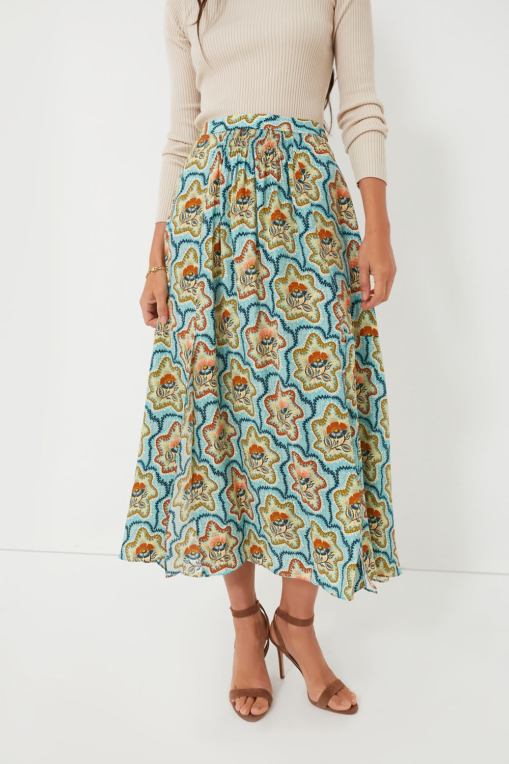 A woman wears a beige long-sleeve ribbed top, the Hunter Bell Tea Time Flower Tucker Skirt featuring a vibrant floral print, and brown strappy sandals.