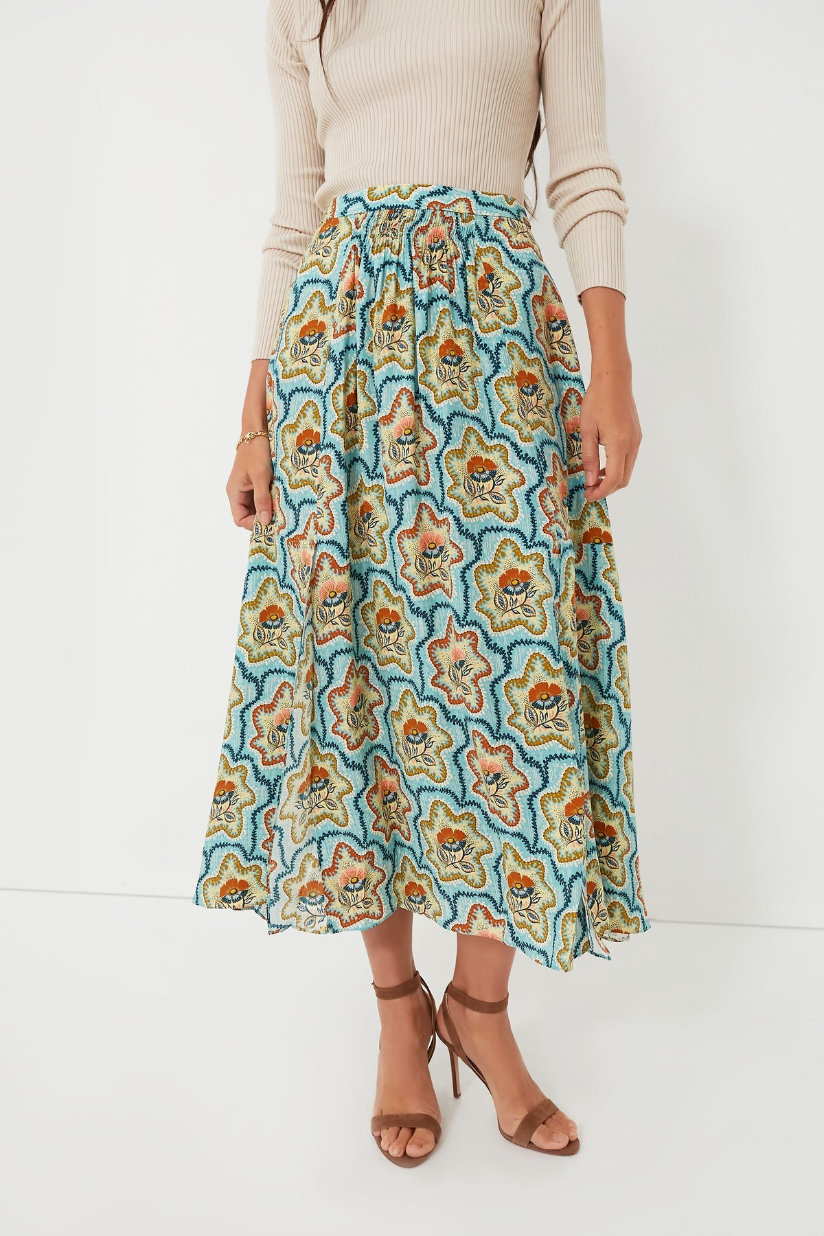 A woman wears a beige long-sleeve ribbed top, the Hunter Bell Tea Time Flower Tucker Skirt featuring a vibrant floral print, and brown strappy sandals.