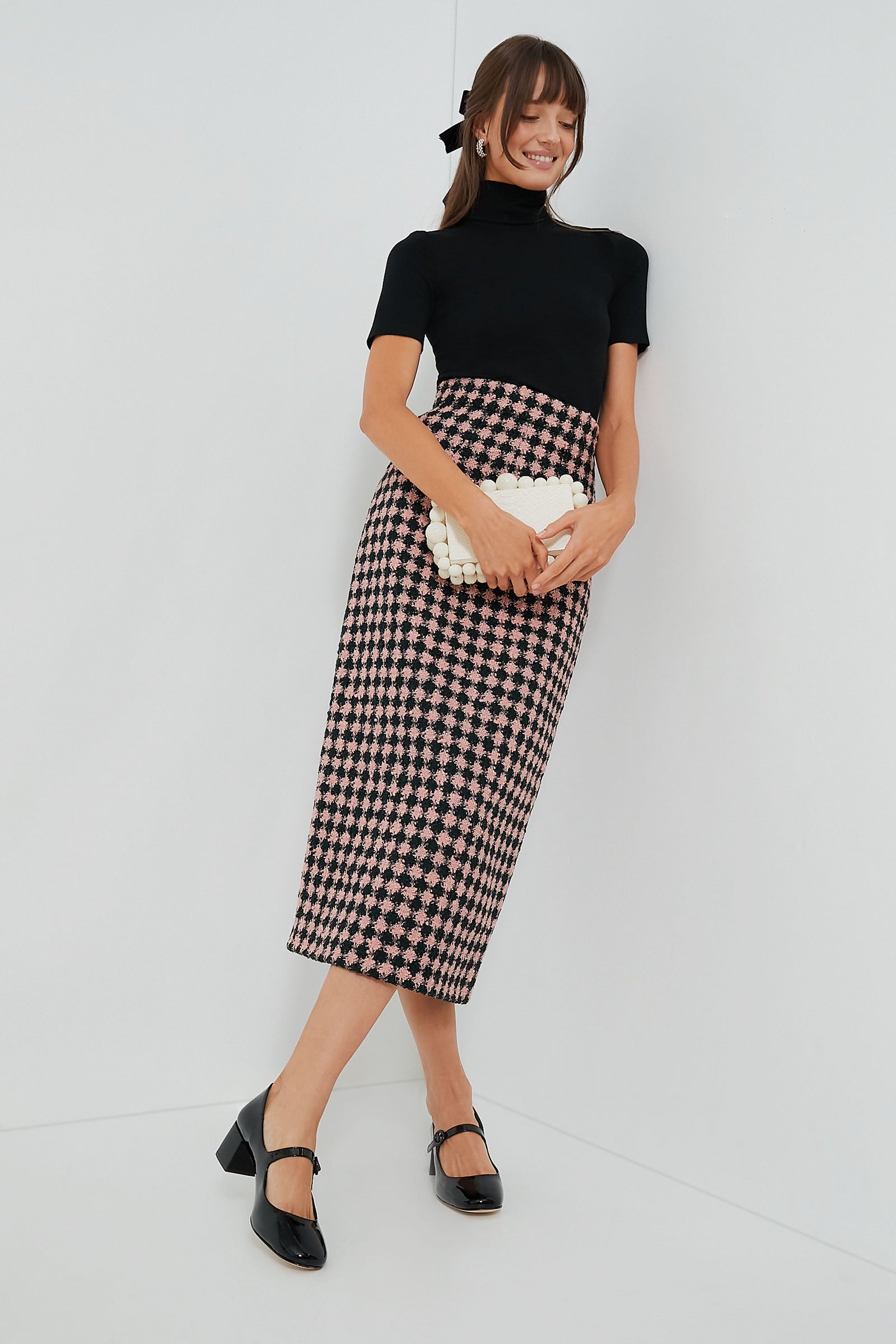 A woman stands against a white wall, styled with a black top, high-waisted pink and black houndstooth skirt, white clutch, black heels, and completes her outfit with a chic Tuckernuck Accessories velvet hair bow.