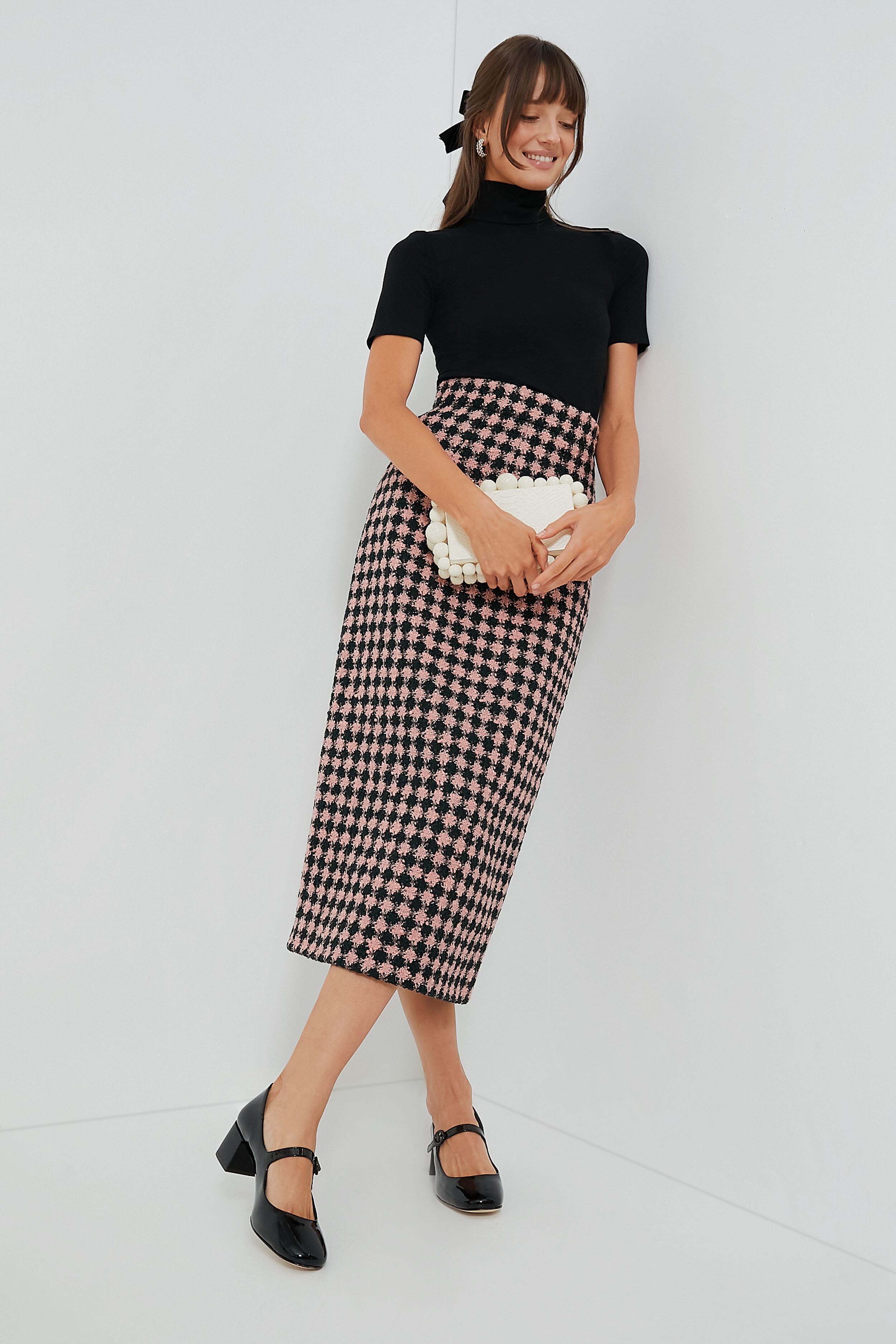 A woman stands against a white wall, styled with a black top, high-waisted pink and black houndstooth skirt, white clutch, black heels, and completes her outfit with a chic Tuckernuck Accessories velvet hair bow.