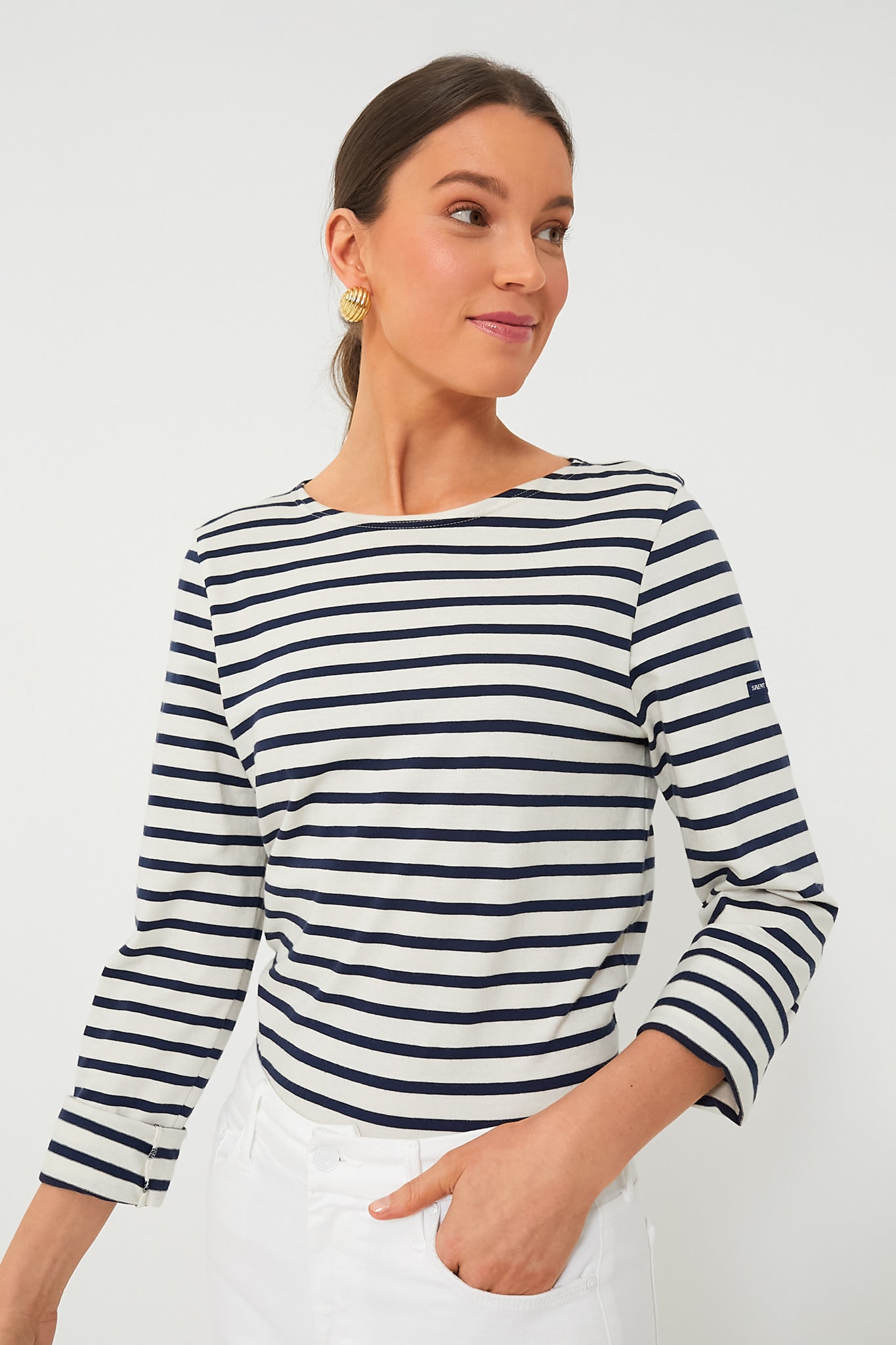 A woman wears a Saint James navy and white sailor stripe long-sleeve tee with white pants, standing in a simple setting.