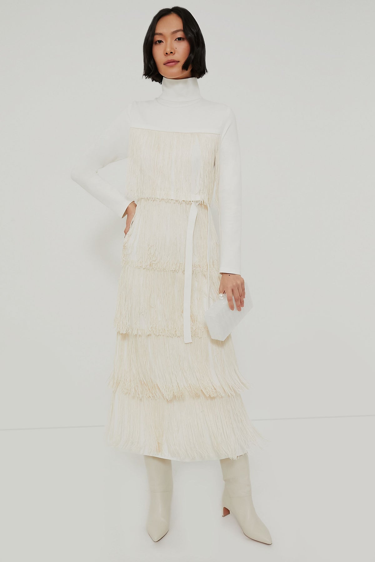 A woman wears an Emerson Fry Ivory Fringe Midi Dress with white knee-high boots and a small white bag for a chic, western-inspired look.
