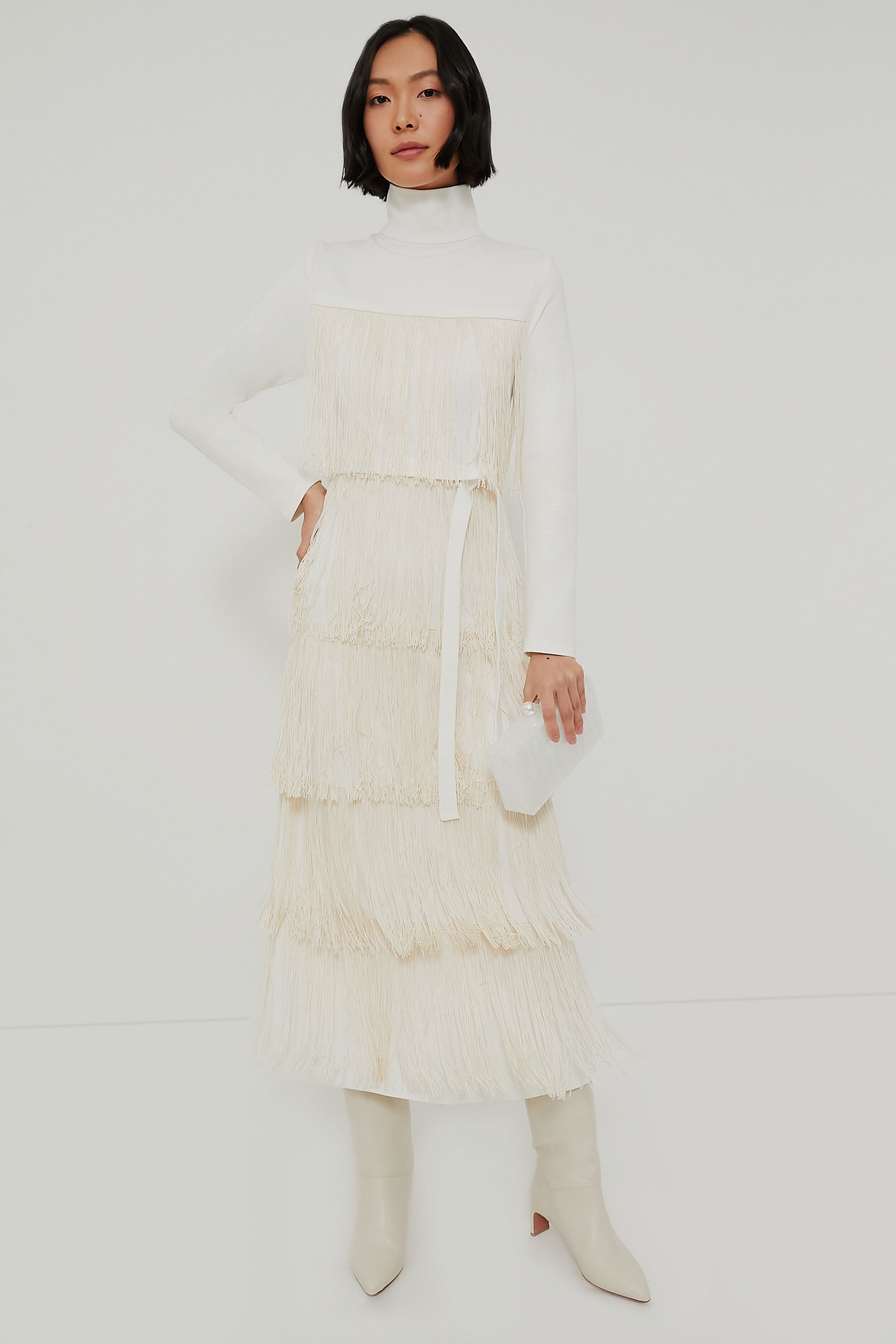 A woman wears an Emerson Fry Ivory Fringe Midi Dress with white knee-high boots and a small white bag for a chic, western-inspired look.